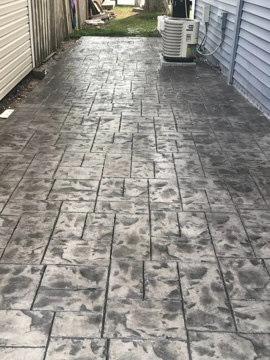 A freshly stamped concrete backyard patio with a gray stone pattern, bordered by a beige house and a blue house, with a grass patch and trees in the background, and an air conditioning unit on the right side.