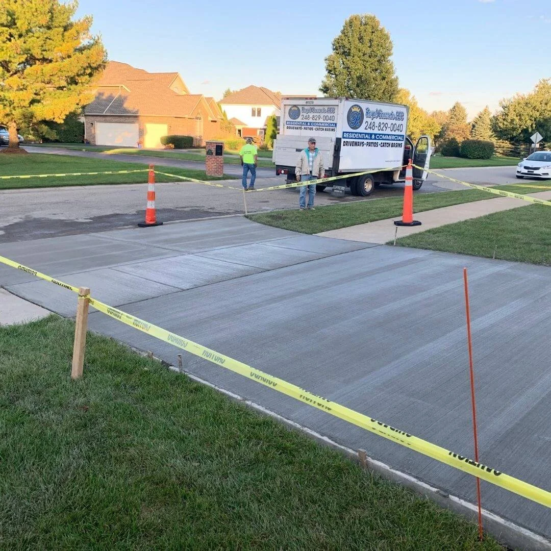 Residential driveway being freshly paved, fenced off with yellow caution tape and orange cones, with a paving truck and two workers present in a suburban neighborhood.