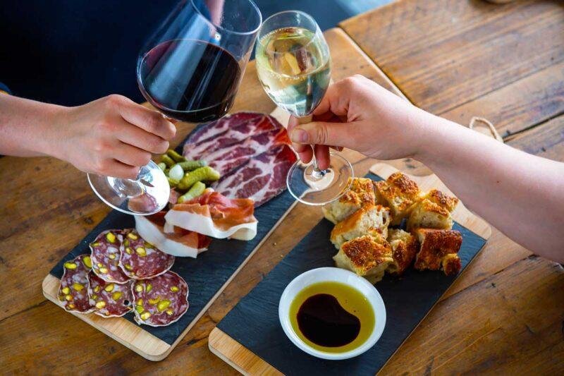 Two people clinking glasses of red wine and white wine over a wooden table with charcuterie and fried food.