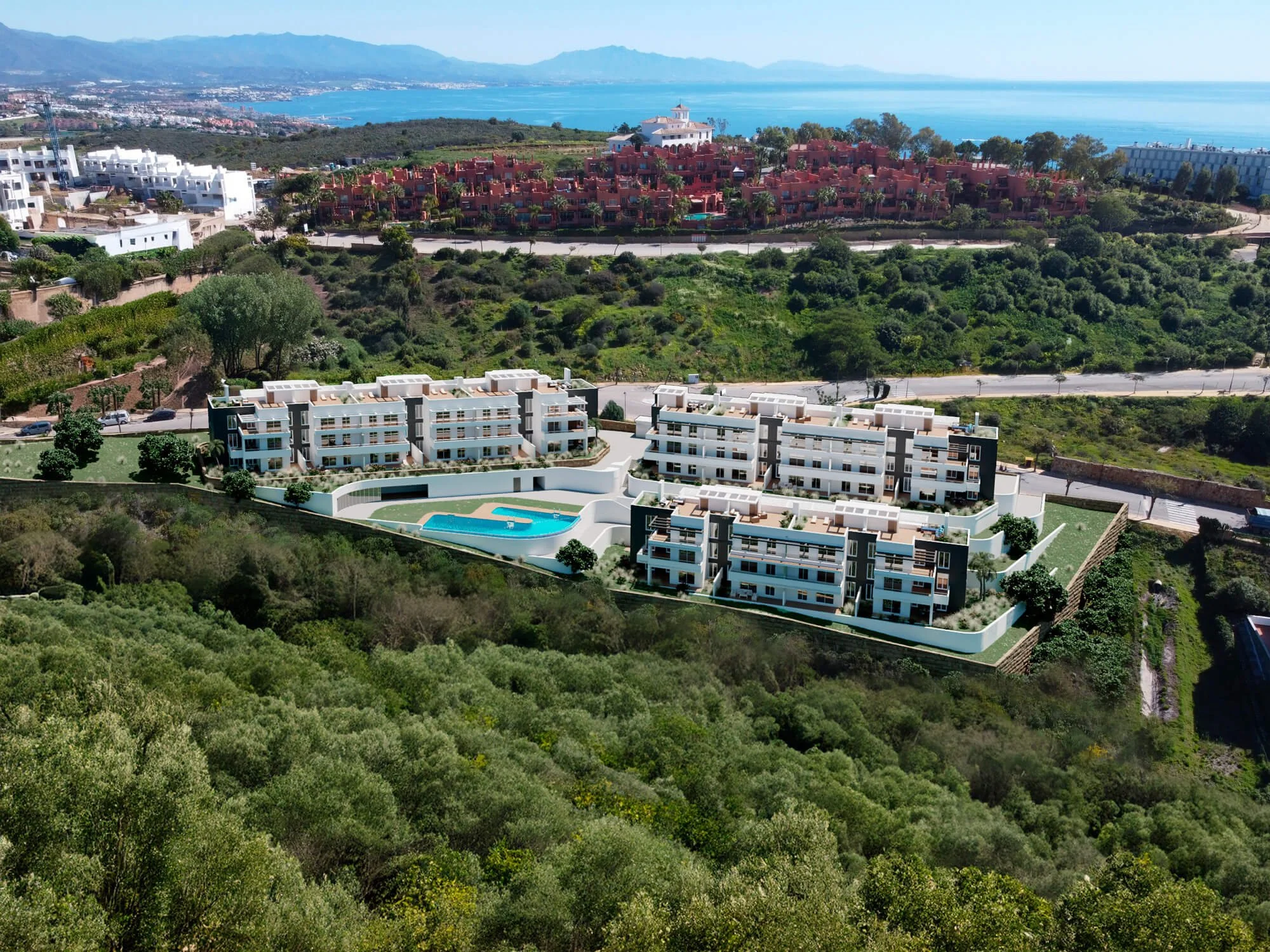 Aerial view of a residential area with modern white apartment buildings, a swimming pool, and roads surrounded by lush green hills, overlooking the ocean and distant mountains under a clear blue sky.