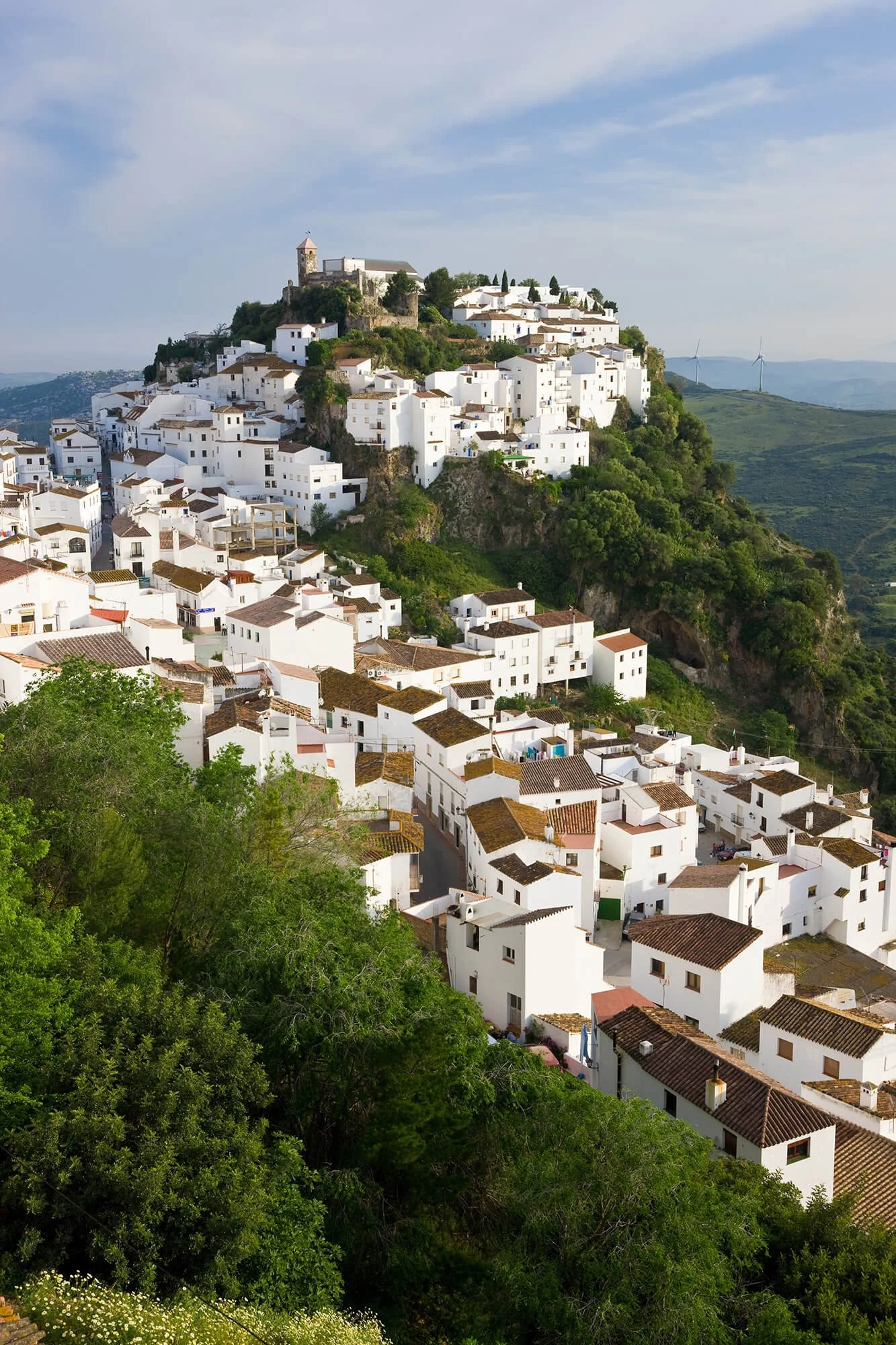 A hillside town with white Mediterranean-style buildings, a castle on top, and wind turbines in the distance under a partly cloudy sky.