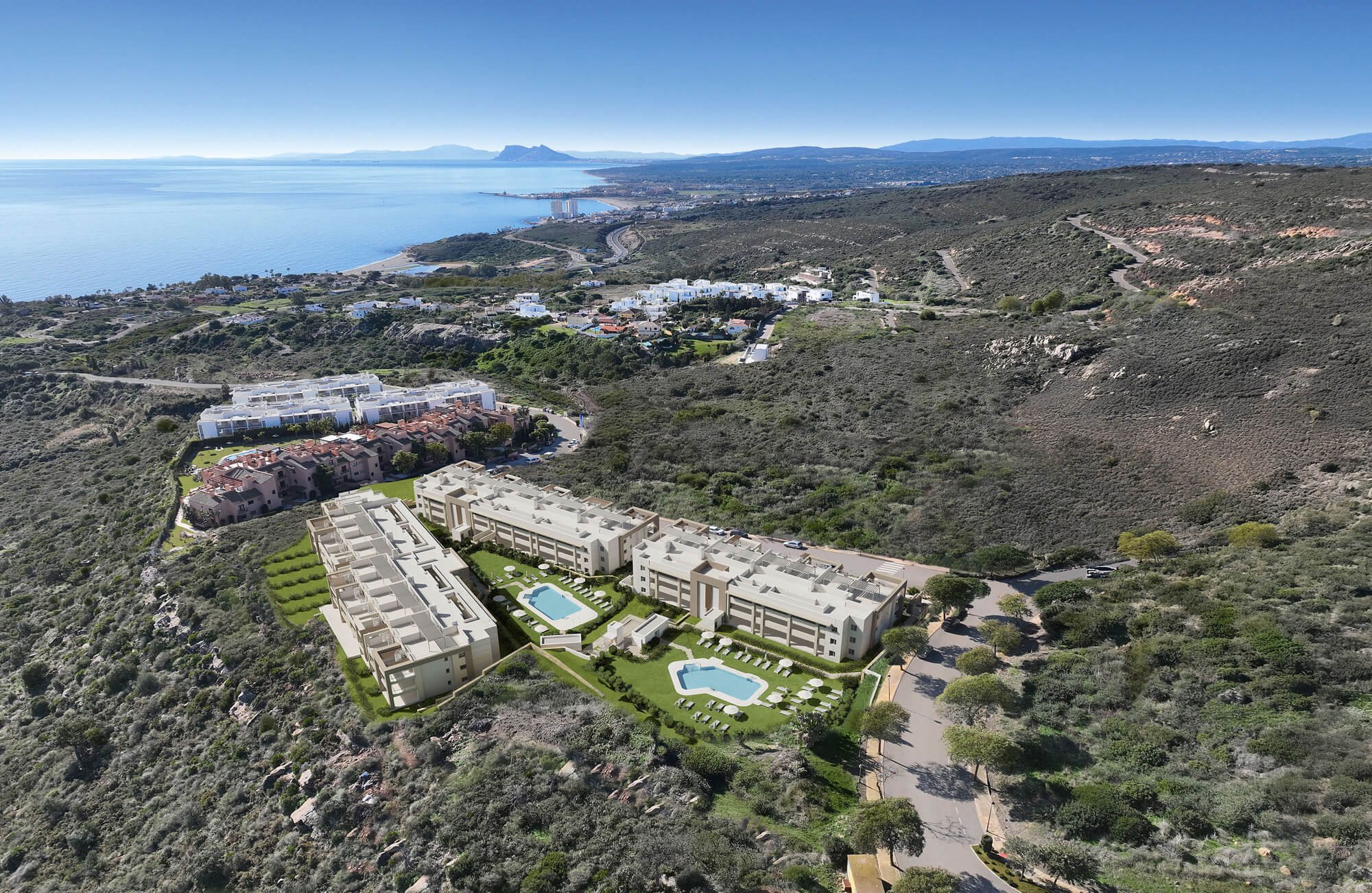 Aerial view of a coastal landscape with modern apartment buildings and swimming pools in the foreground, hilly terrain with sparse vegetation, a shoreline with a few buildings, and distant mountains under a clear blue sky.