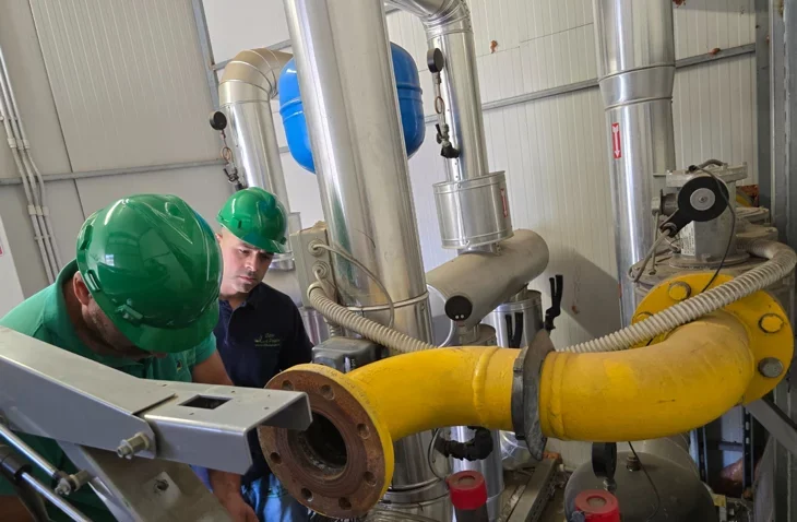 Due operai con caschi verdi lavorano in un impianto industriale con tubi e macchinari metallici.
