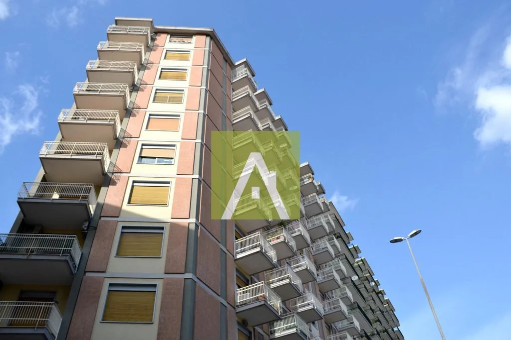 Edificio residenziale con più piani e balconi, sotto un cielo blu con alcune nuvole sparse, e una lampada pubblica in strada.