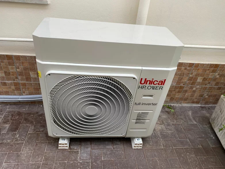 Un'unità esterna di aria condizionata con marca Unical HP Power, full inverter, installata su un balcone di fronte a una parete con piastrelle marroni e un bordo bianco, con tubi e cavi collegati.