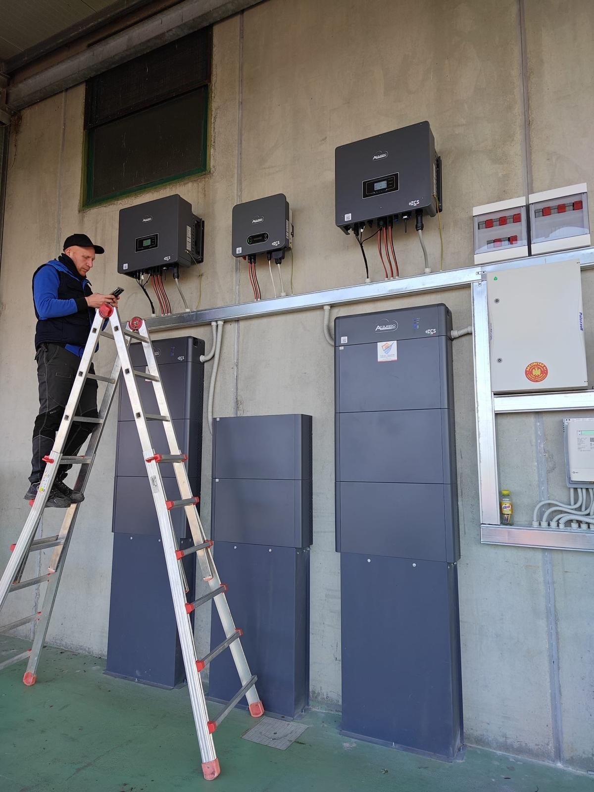 Un uomo con una scala sta lavorando con apparecchiature elettroniche e pannelli di controllo montati su una parete in un ambiente industriale o di servizio.