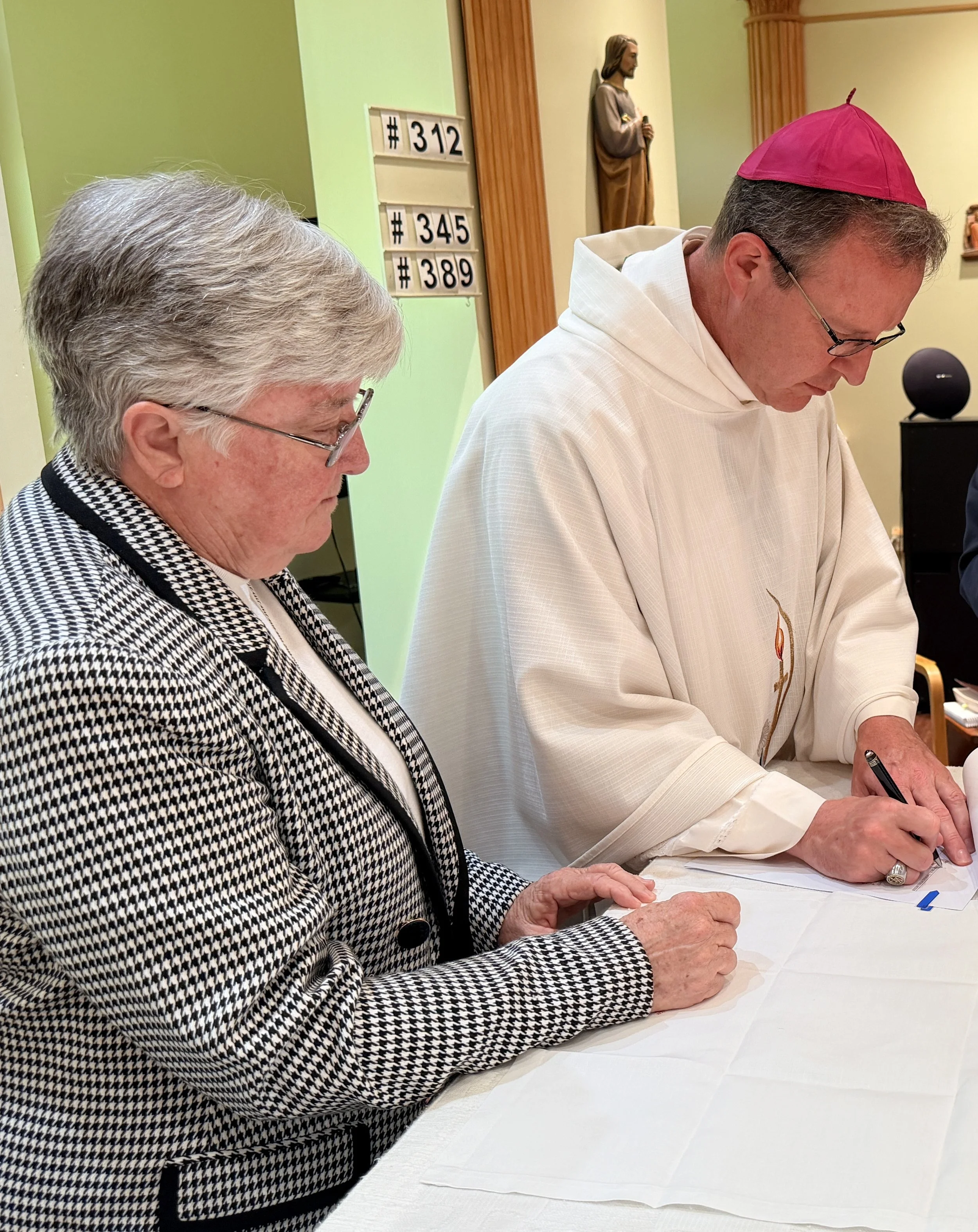 A priest in white robes and a pink skullcap signing a document with a woman in glasses and a checkered jacket also signing a document at a table, with religious statues and signs with numbers in the background.