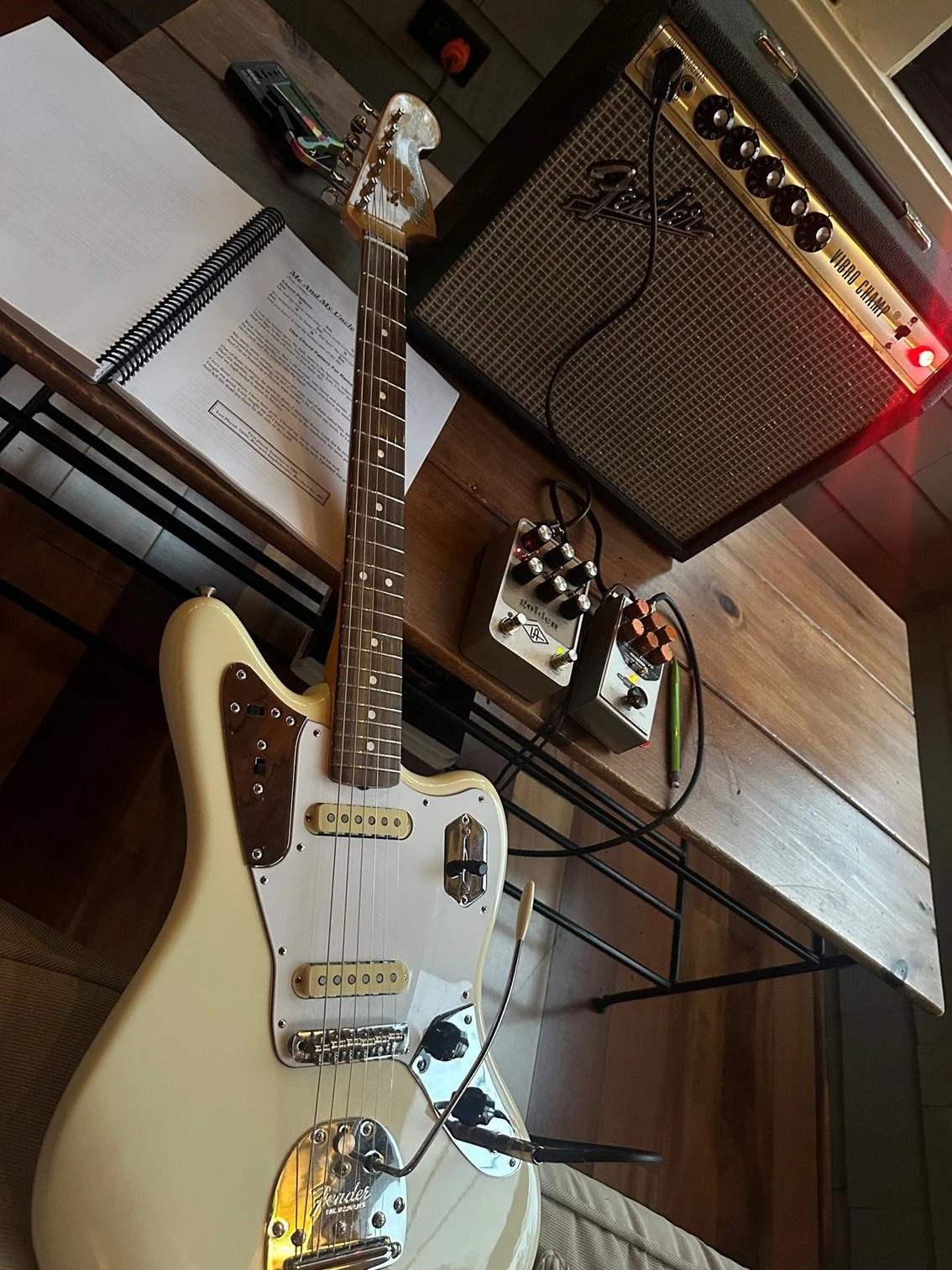 An electric guitar lying on a table, connected to guitar pedals and an amplifier, with an open book and a notebook nearby.