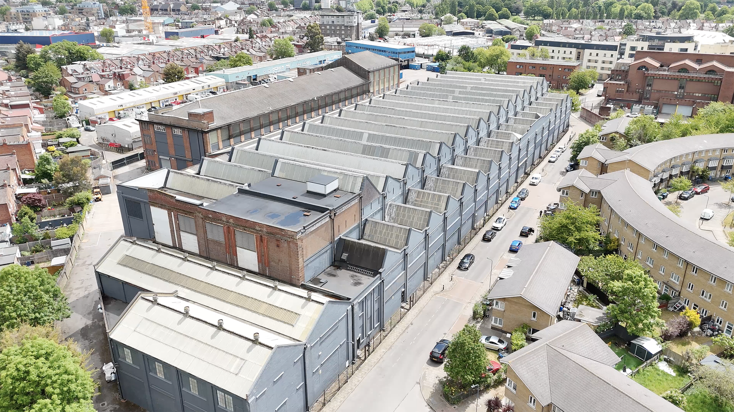 Aerial view of an urban area with a mix of residential and industrial buildings, including a large factory with a series of angular, sloped rooftops and a parking lot with cars nearby, surrounded by trees and other housing structures.