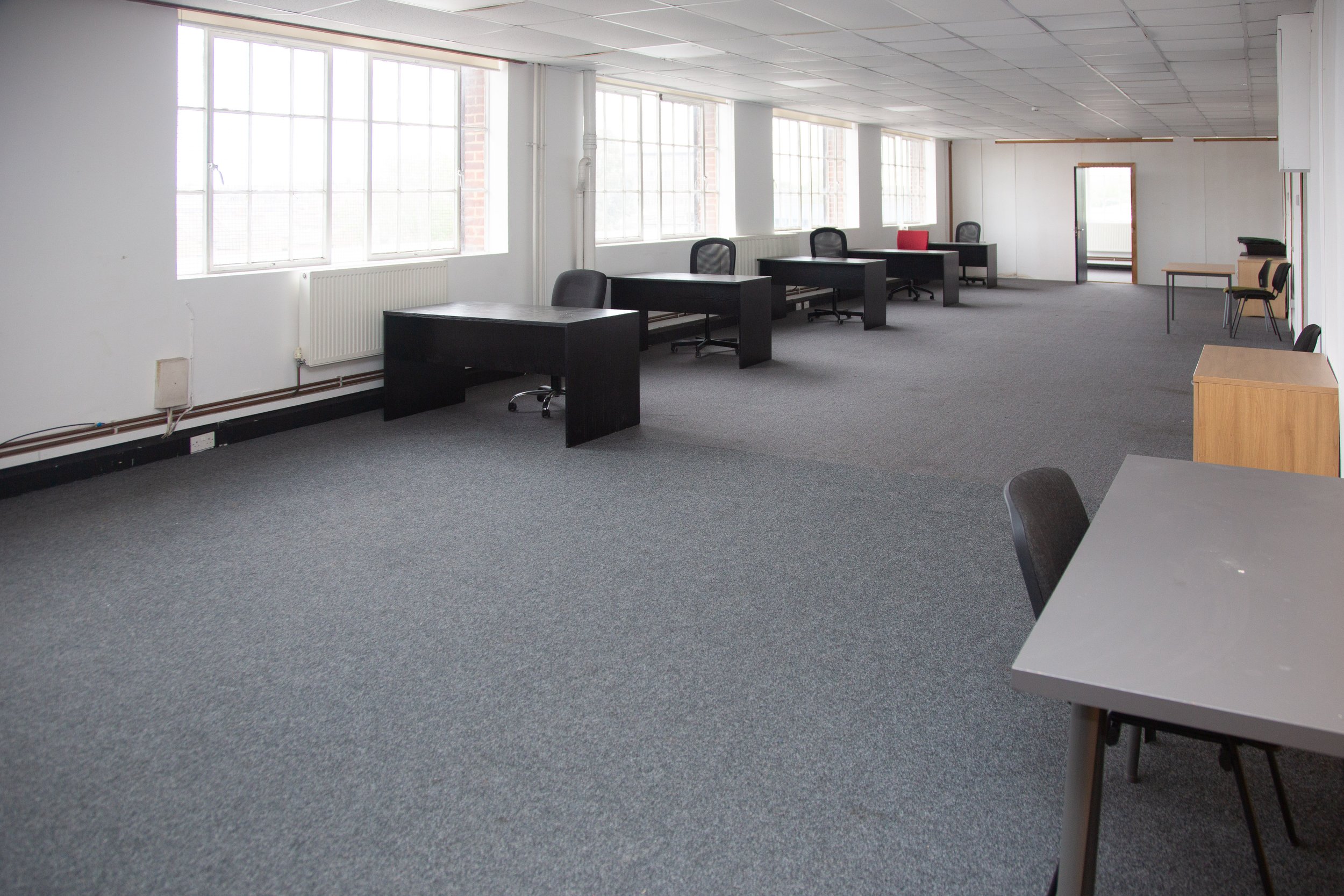 An empty office space with large windows, black desks, office chairs, and a gray carpeted floor.