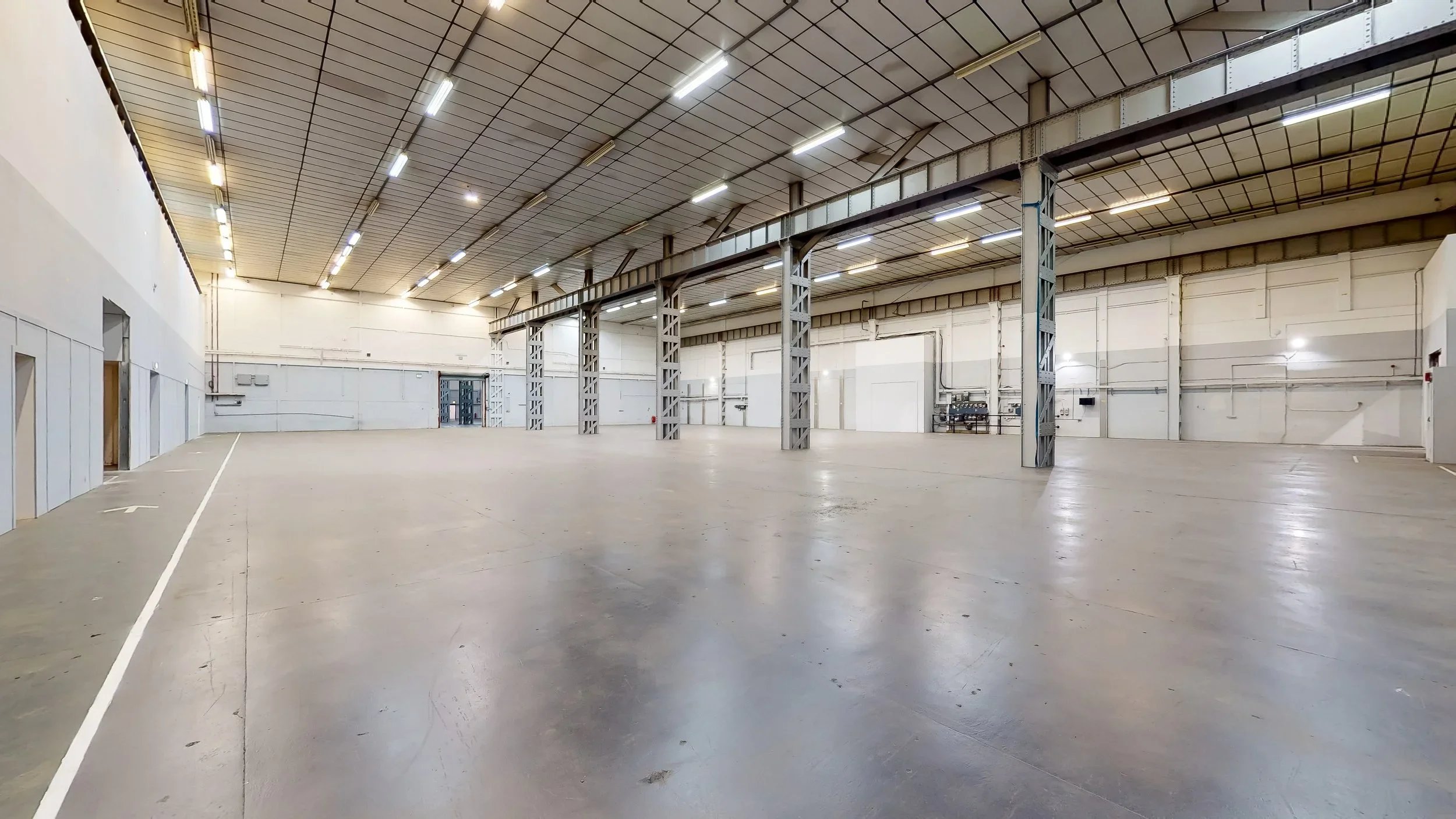 Empty warehouse with high ceilings, metal support beams, white walls, and bright overhead lighting