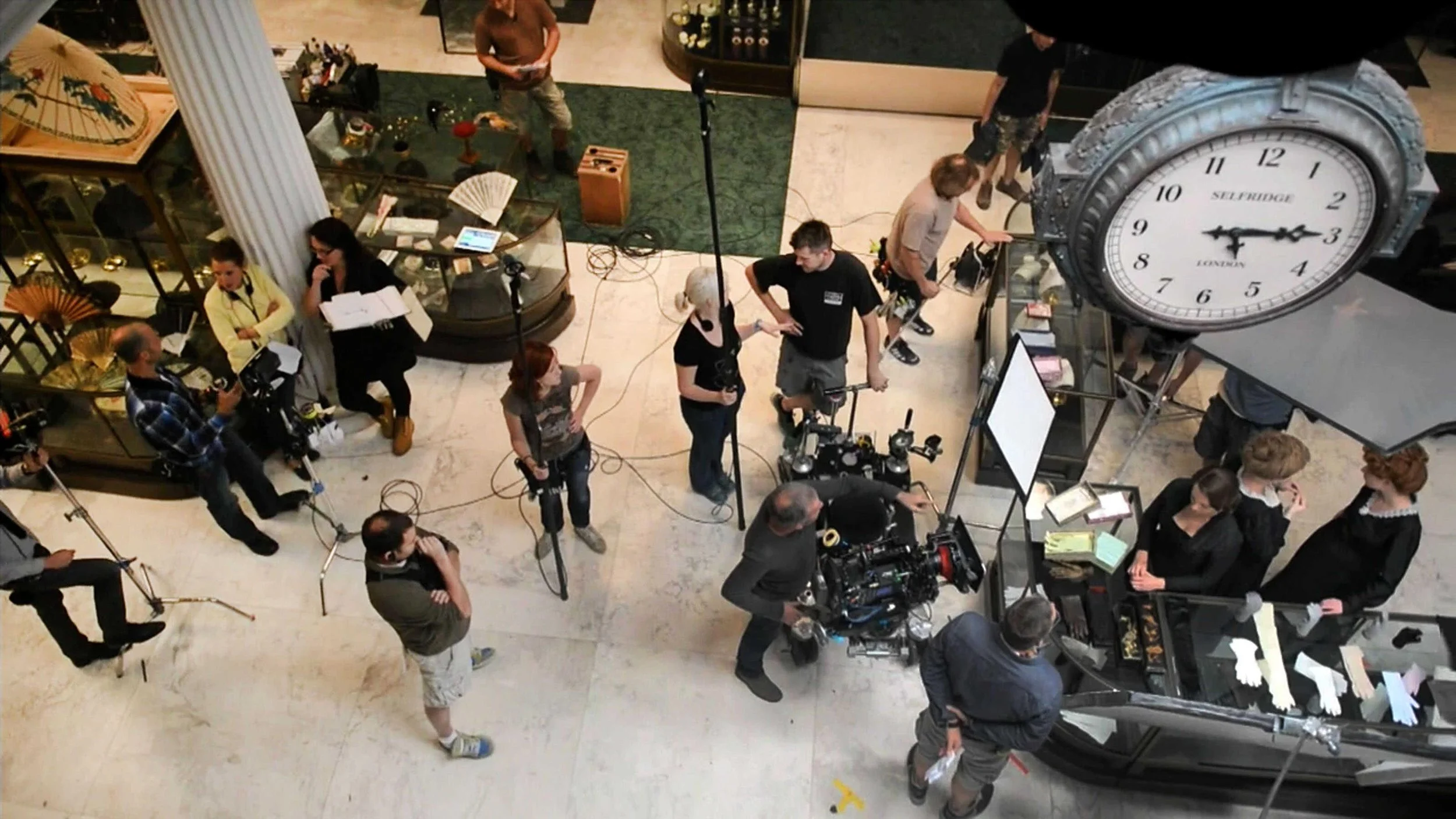 A film crew sets up equipment on the lobby of a building. Several people are working with cameras, lights, and other filming gear, and a large clock displaying the time is visible in the scene.