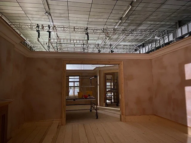 Interior view of a building under construction with unfinished walls and an exposed ceiling with scaffolding and lighting equipment.