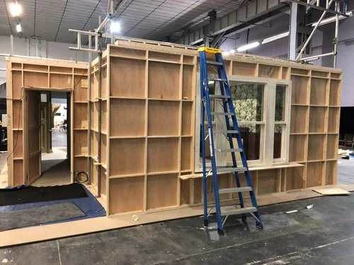 Wooden framing of a small building or room under construction with a blue ladder leaning against it inside a warehouse or workshop.