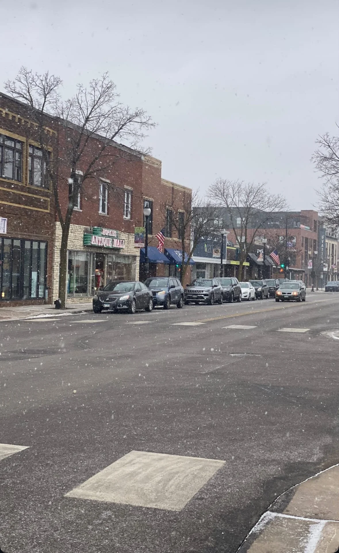 A street scene with parked cars along a row of brick buildings on a snowy day, with leafless trees and American flags.