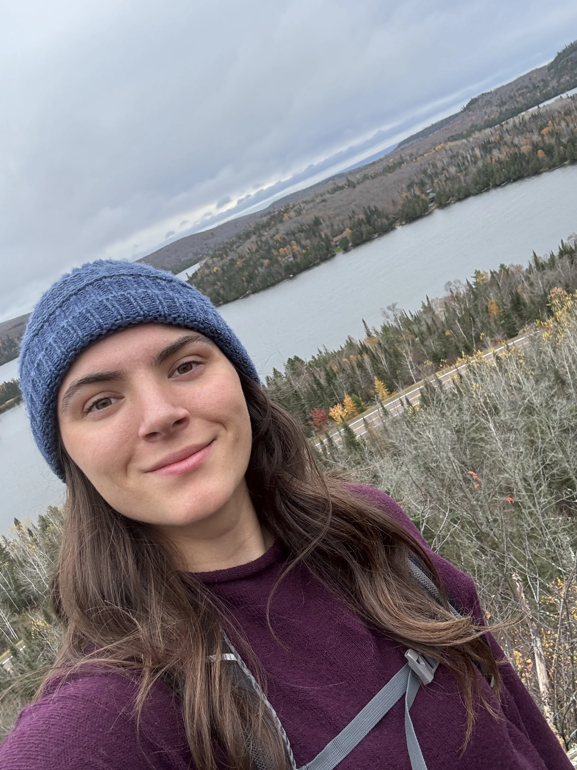 A young woman with long brown hair and fair skin outdoors near a lake, wearing a blue knit beanie and a purple jacket, with a slight smile, during autumn with trees showing fall colors.