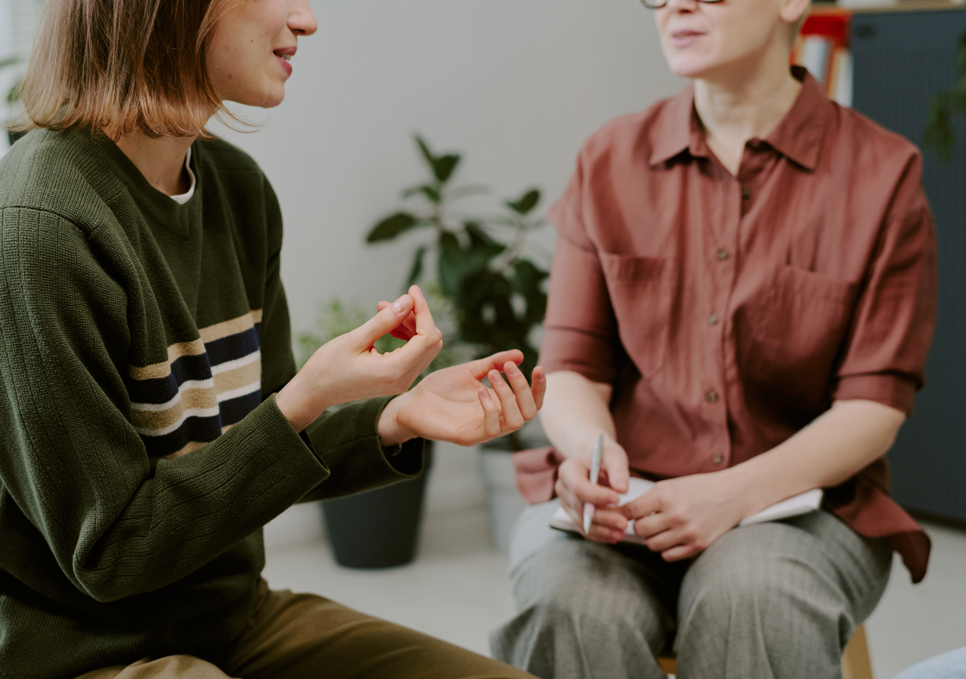 Two women having a conversation, one is showing something on her hand to the other, holding a pen and notebook, in an office setting with a plant and bookshelf in the background.