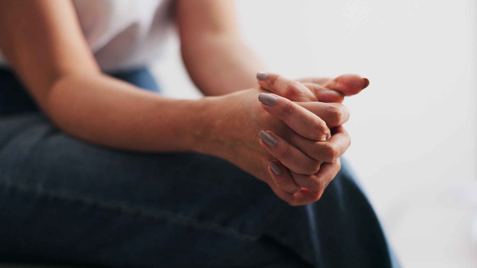 Close-up of a person's hands resting on their lap with fingers gently intertwined, wearing a white shirt and blue jeans, against a plain white background.