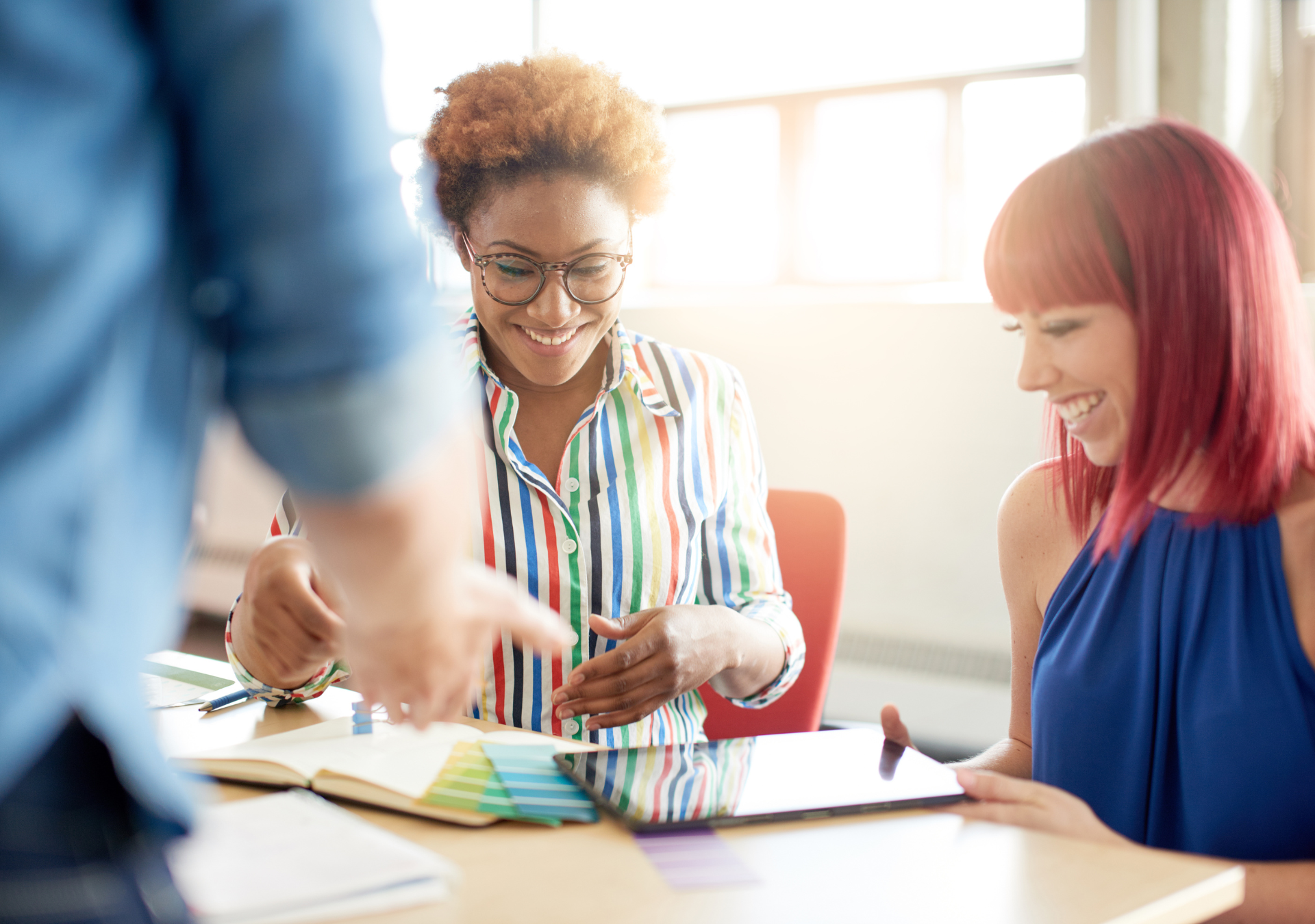 Two women smiling and looking at color samples or fabric swatches on a table in a well-lit room, with another person partially visible in the foreground.