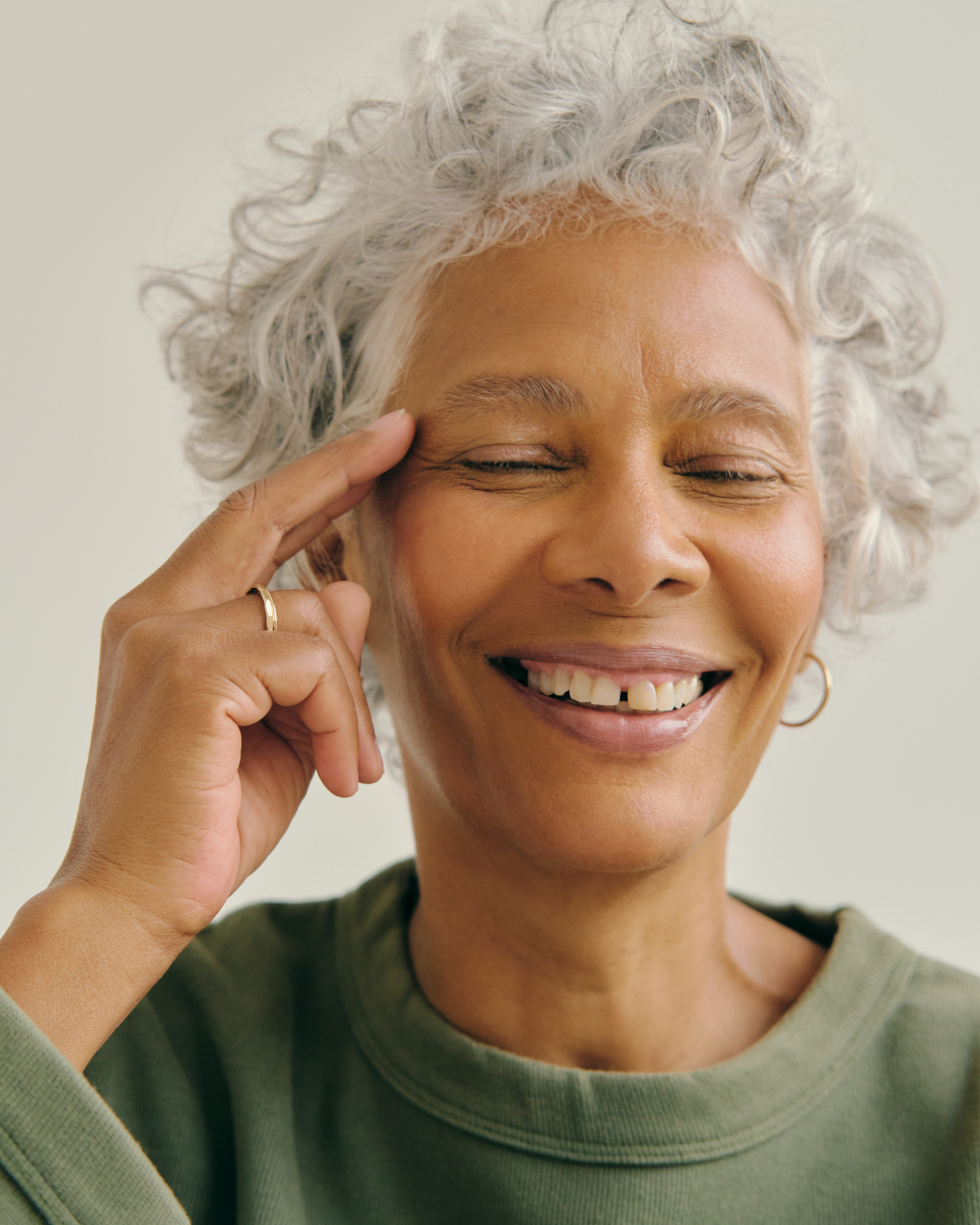 A joyful older woman with short curly gray hair, smiling with eyes closed, touching her temple with her index finger, wearing a green top and hoop earrings.