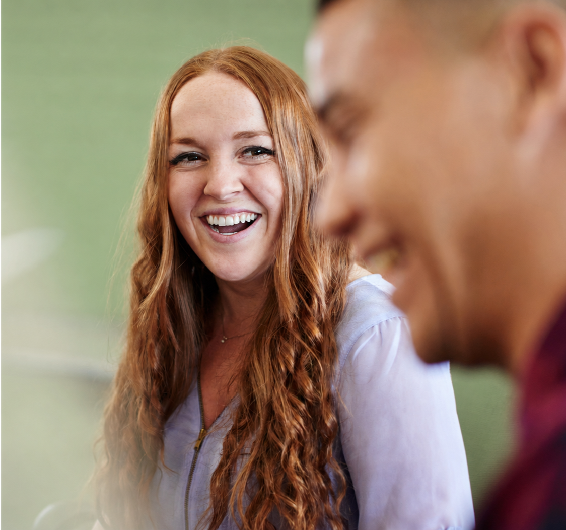 A woman with long, wavy red hair smiling and looking at a man whose face is partially visible in the foreground.