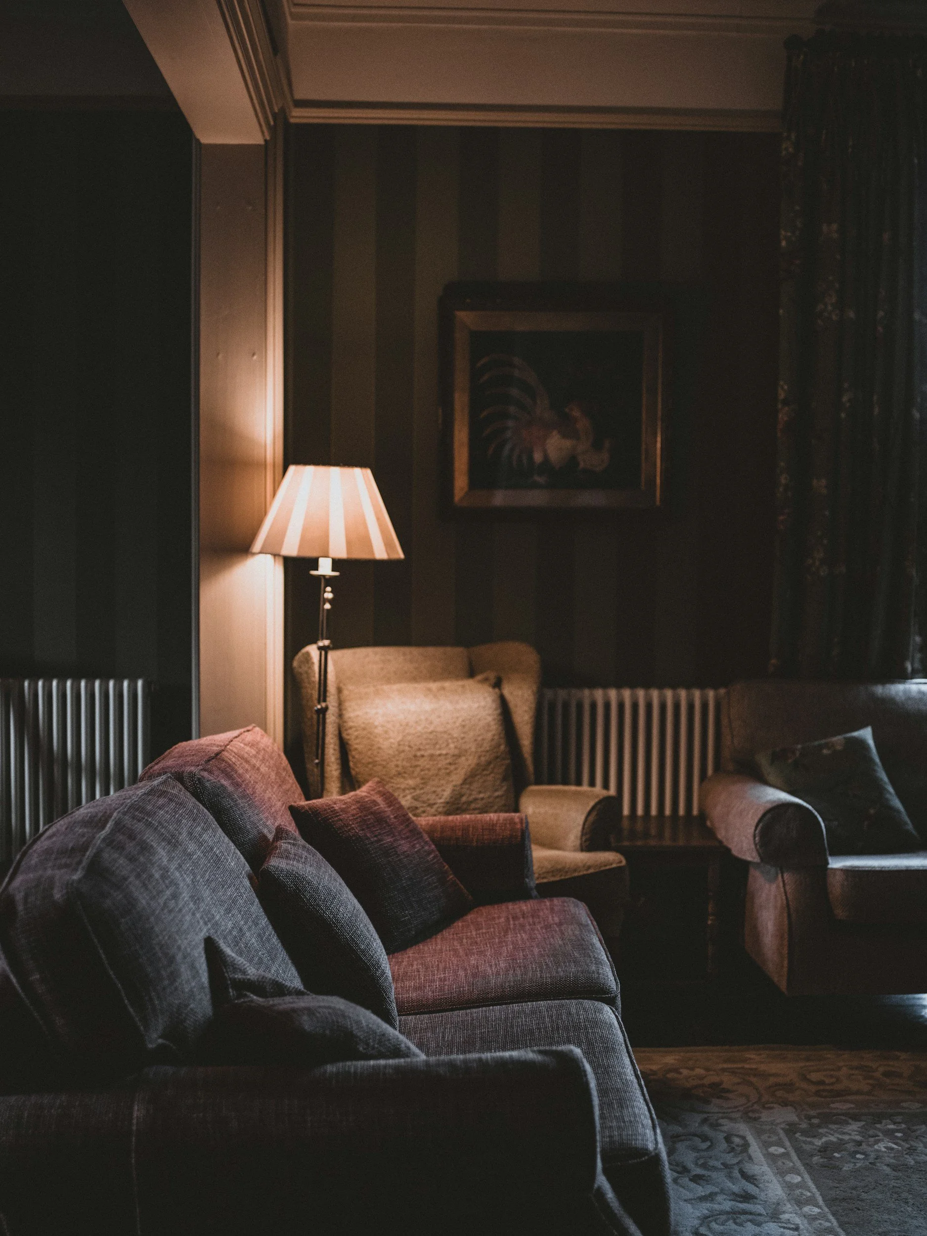 A cozy living room with a patterned sofa and pillows, a side table with a rolled-up blanket, a vintage armchair, a table lamp, a framed rooster painting, and dark curtains, illuminated by warm lighting.