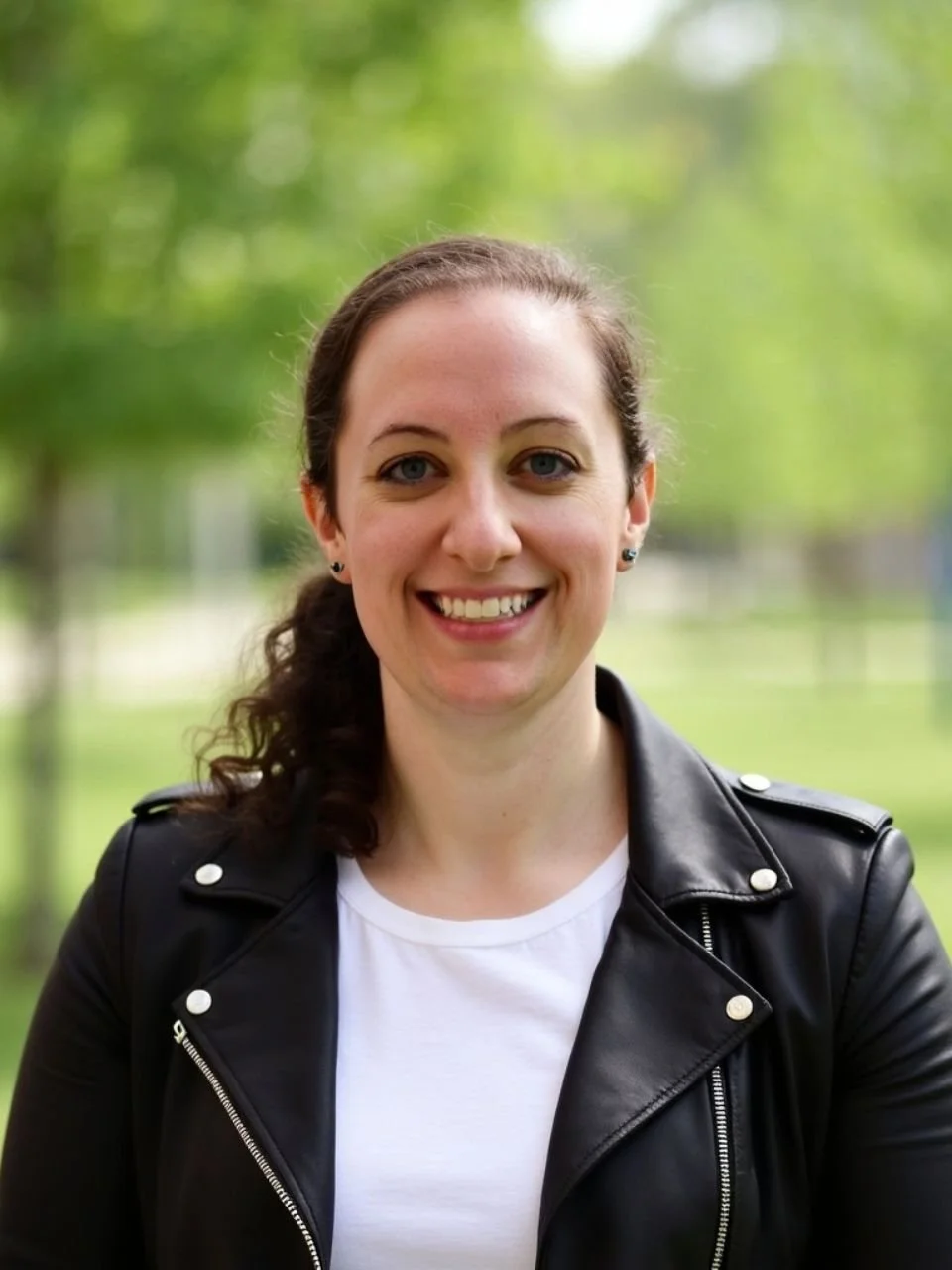 A smiling woman with brown hair and blue eyes, wearing a black leather jacket and white shirt, standing outdoors with blurred green trees in the background.