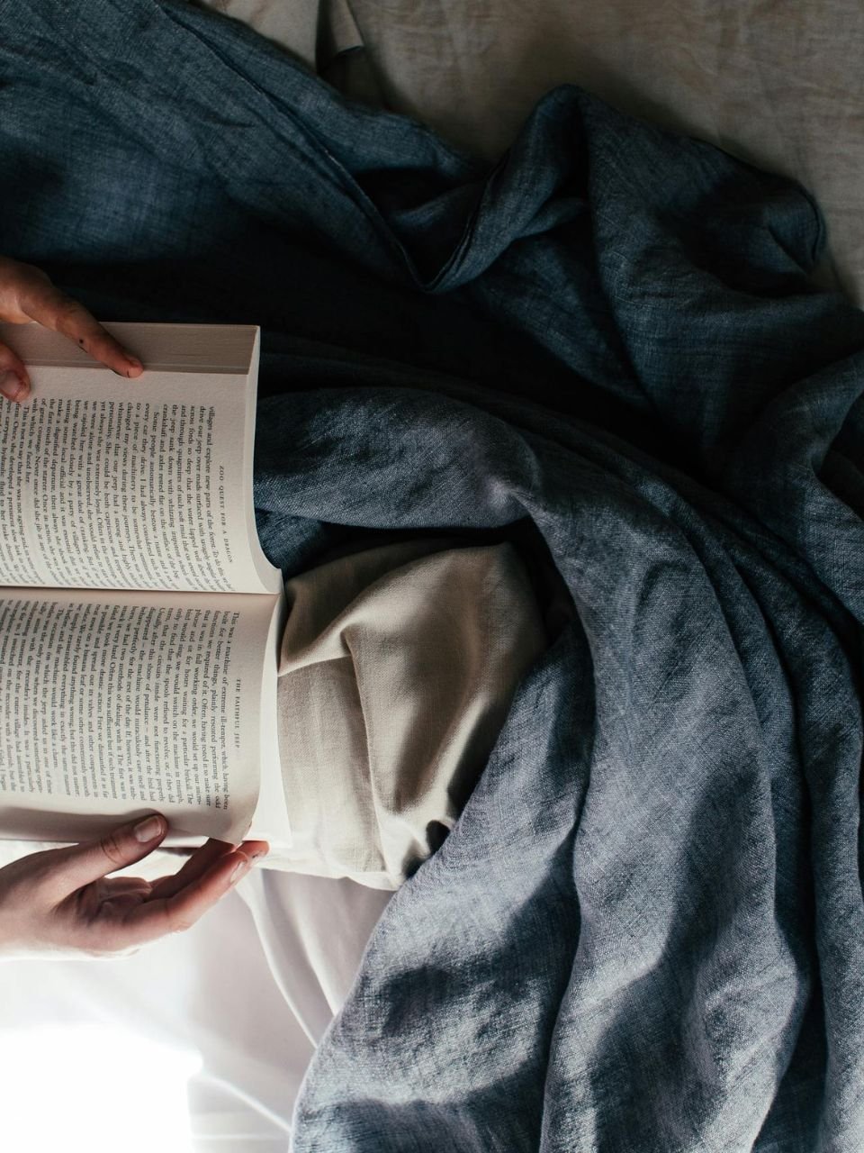 Person reading a book while sitting on a gray couch or bed, wearing blue jeans and gray pants or blanket.