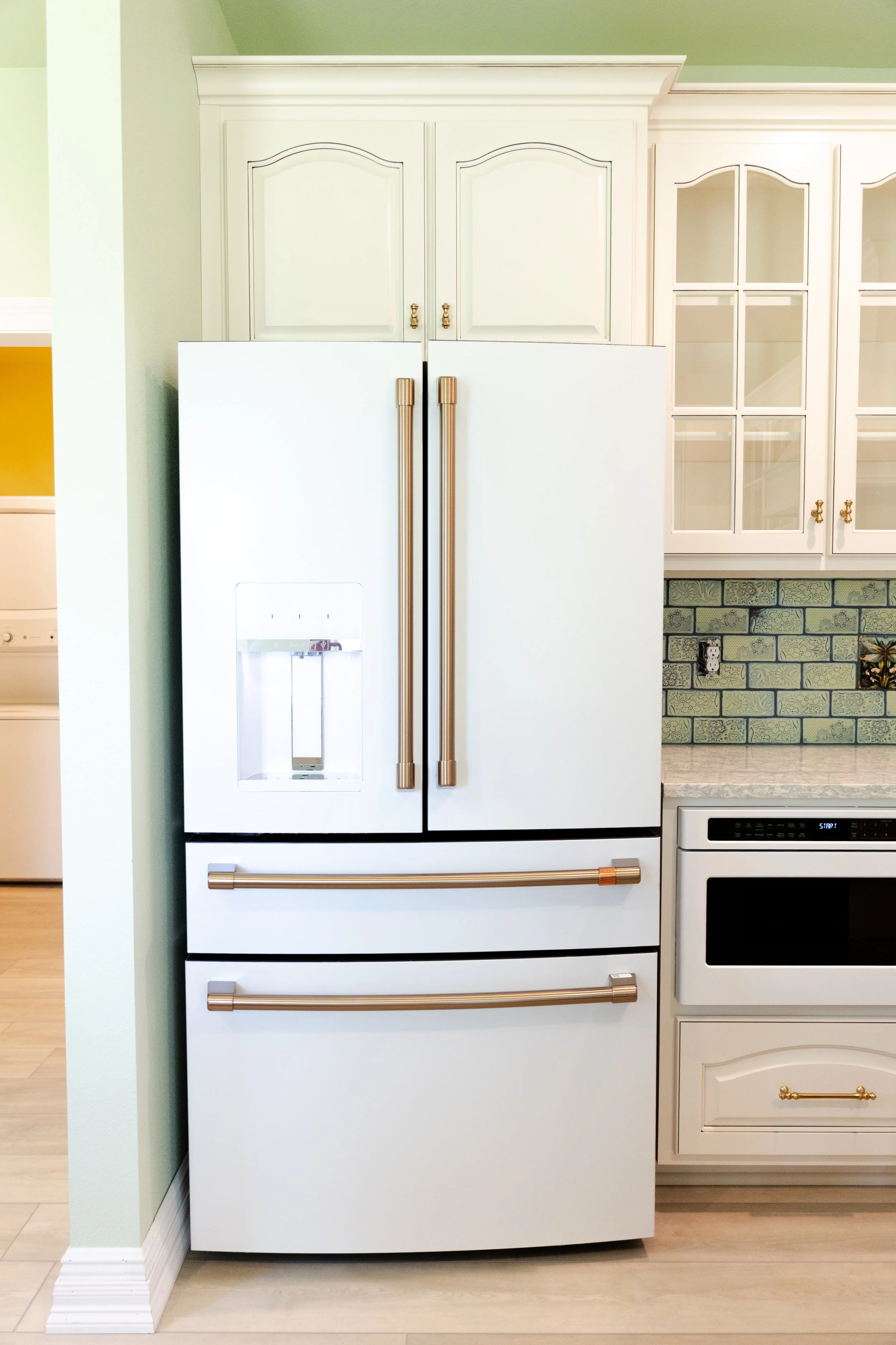 A white refrigerator with gold handles in a kitchen with white cabinetry and patterned green tile backsplash.