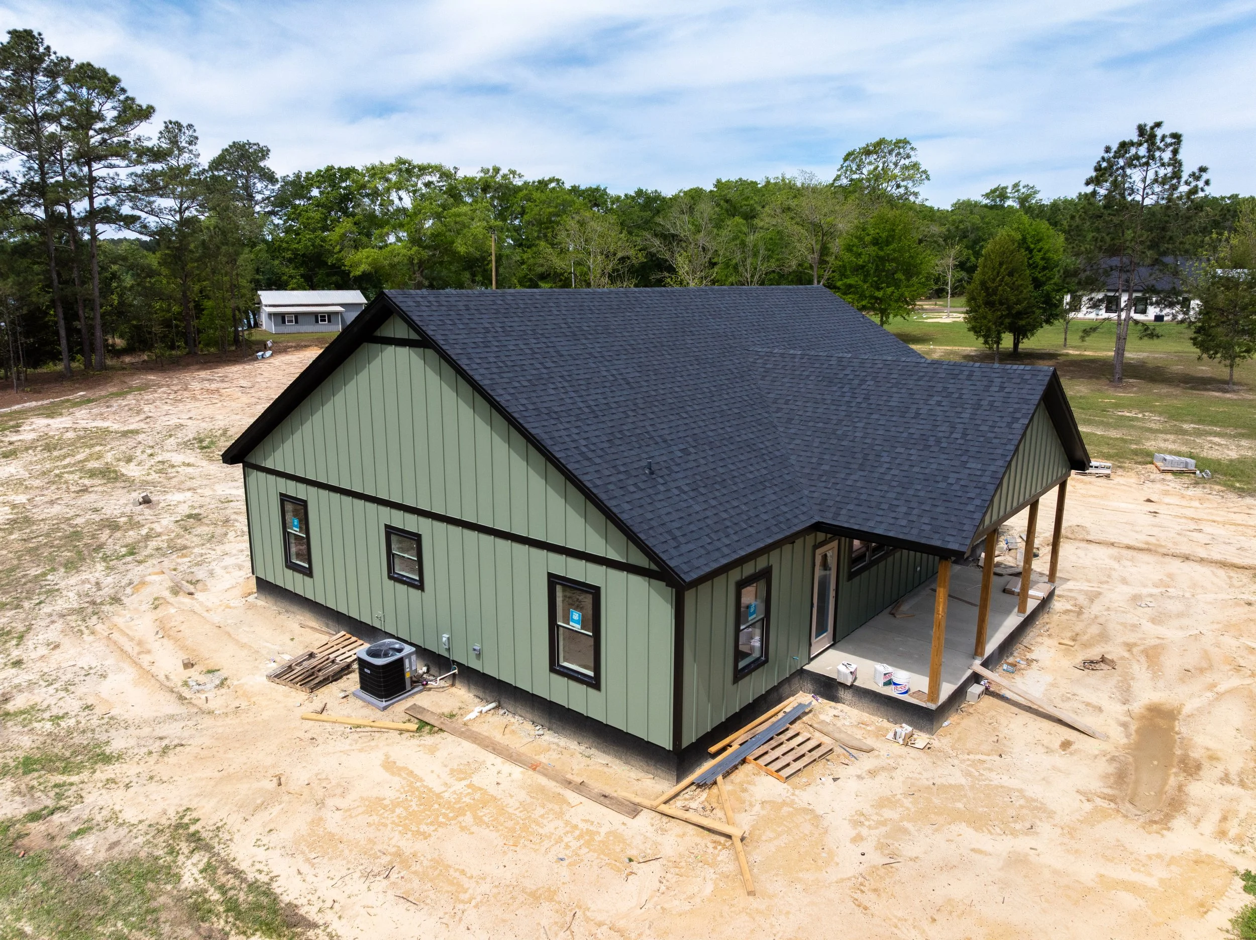 A house under construction with a black roof, tan and black siding, and several tools and construction materials around. There are trees and a blue sky in the background.