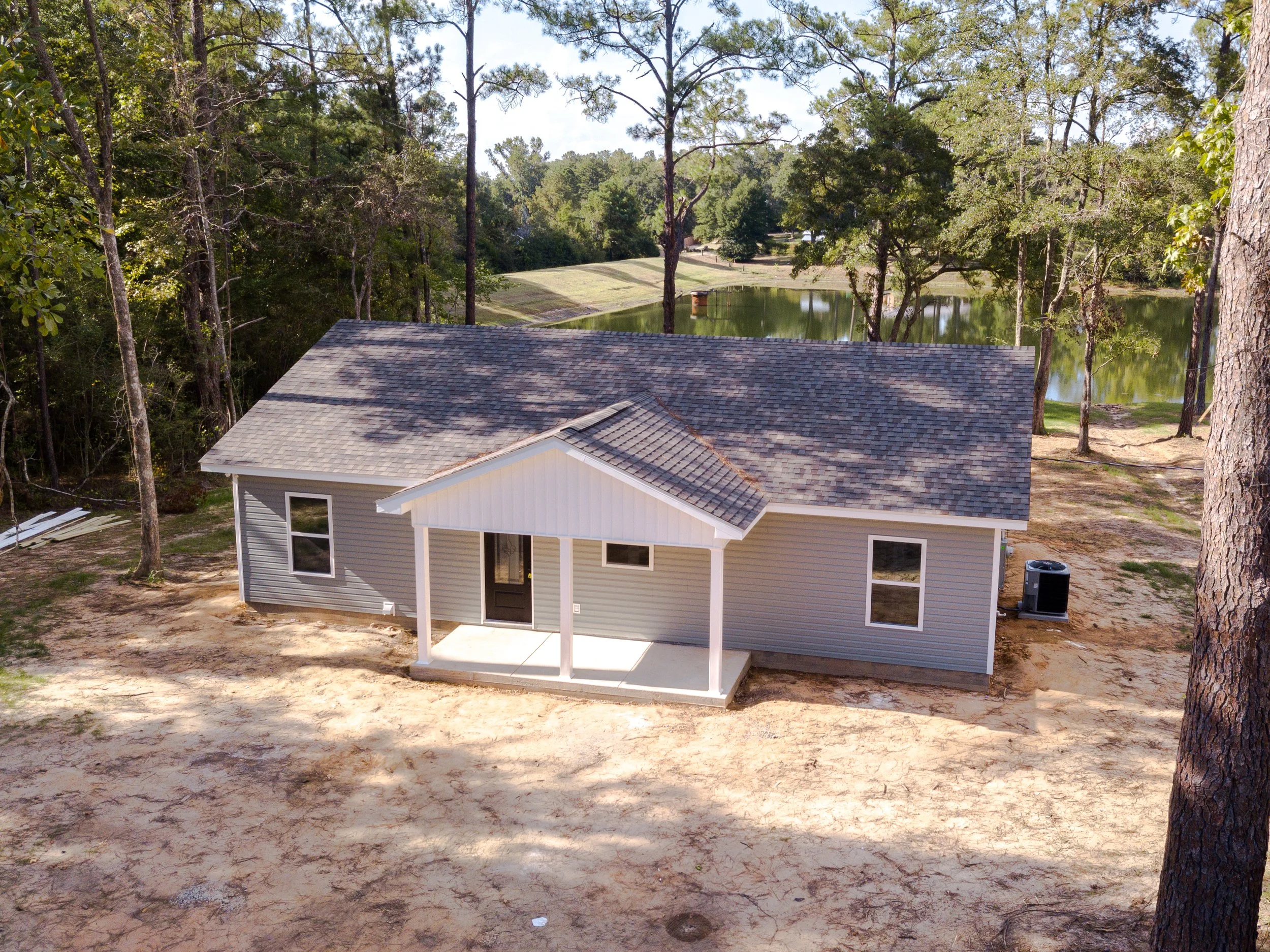 A house under construction with a gray shingle roof, white siding, and a small porch. Surrounding the house are tall trees, a pond in the background, and partly cloudy sky.