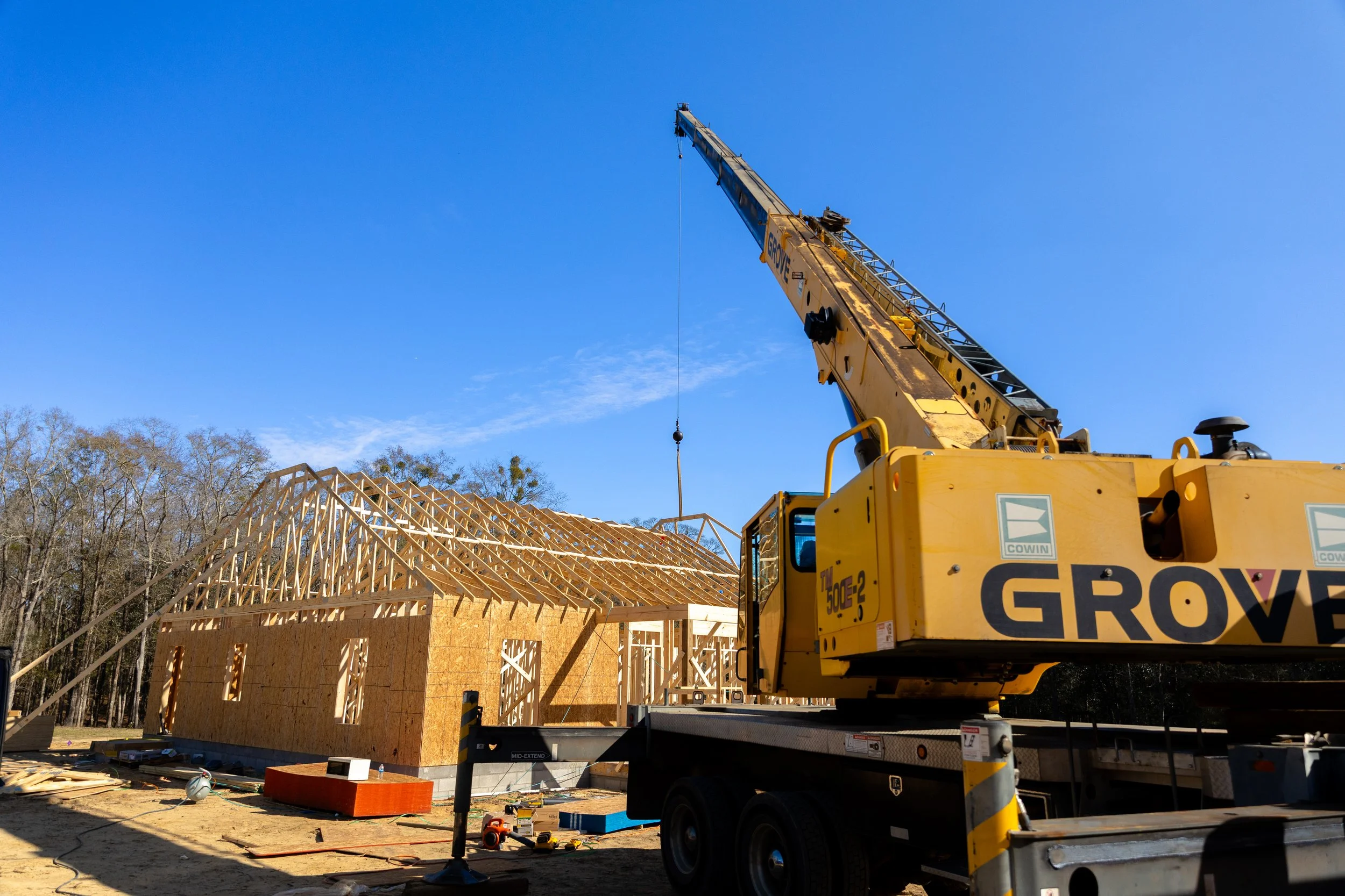 Construction site with wooden framework for a building, a large yellow crane, and construction tools and materials on the ground under a clear blue sky.