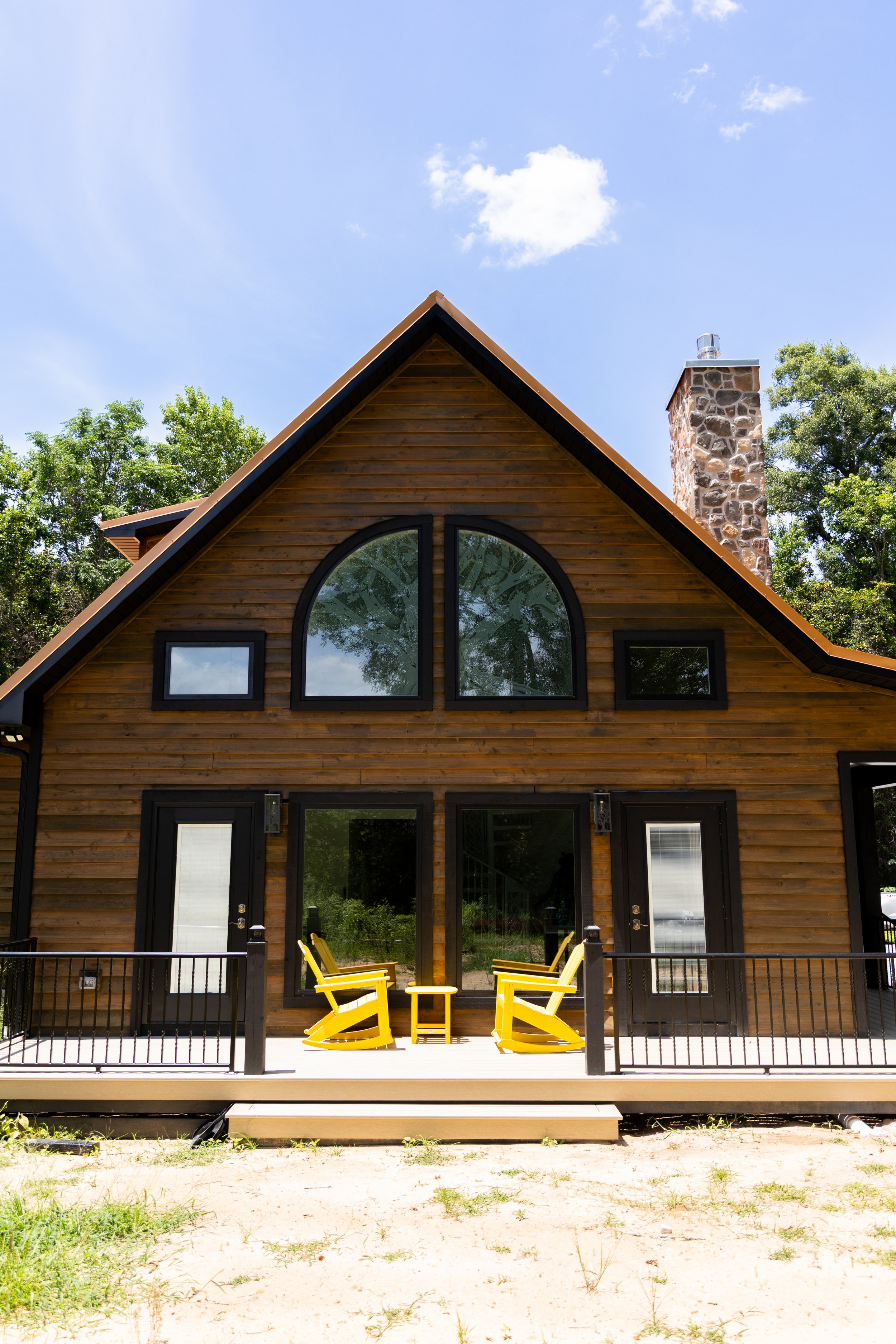 A wooden house with a steep gabled roof, large arched window, and a stone chimney, set against a blue sky with clouds. The house has a balcony with black railing, with yellow outdoor chairs and a small yellow table on it.