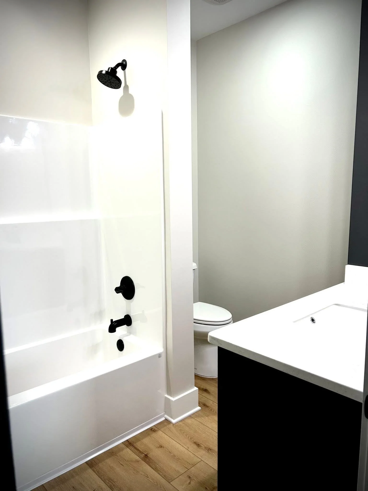 A modern bathroom with a white shower area, black fixtures, a white toilet, a white sink with black accents, and wood flooring.