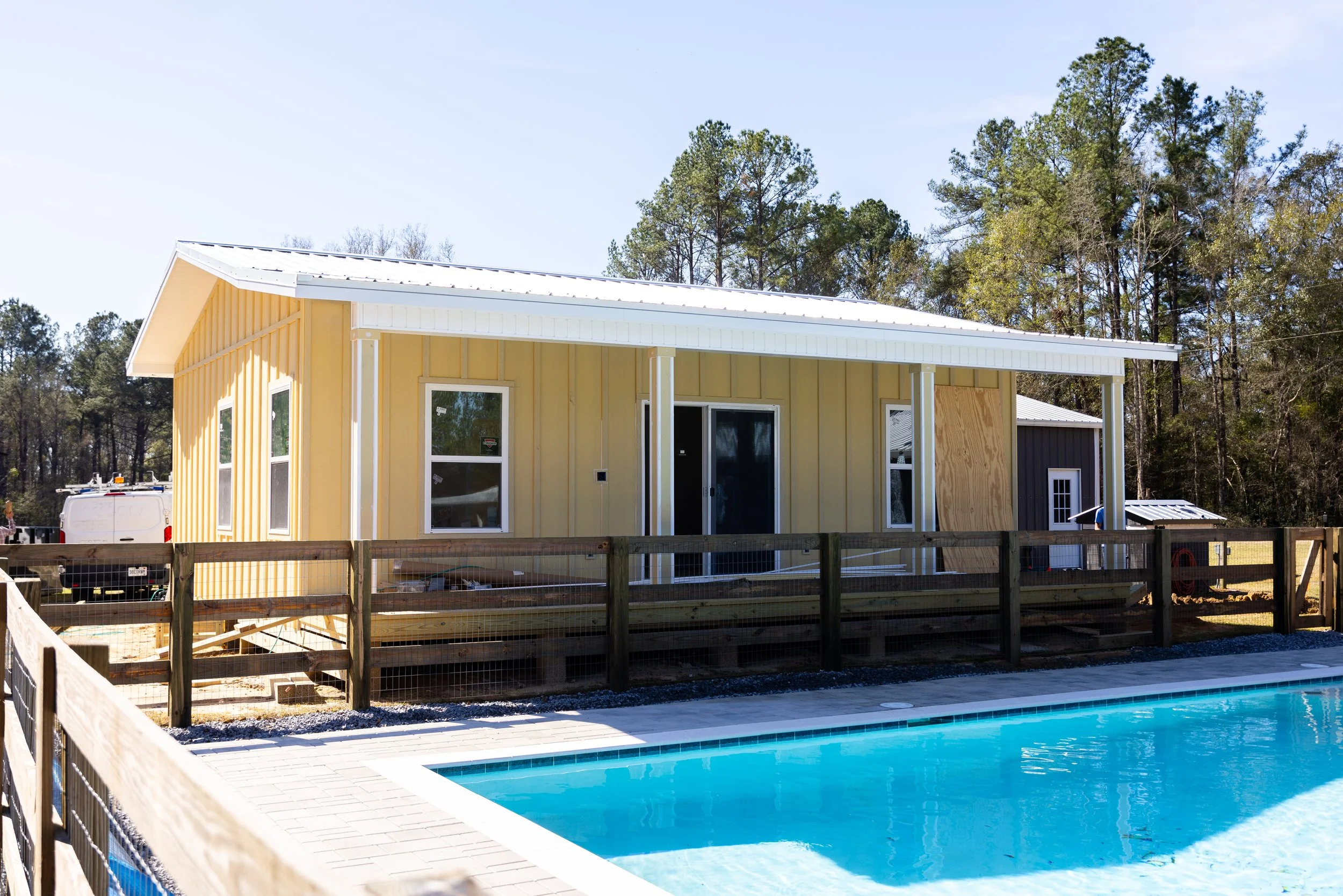 Newly constructed yellow house with large windows and a sliding glass door, under a white metal roof, with a fenced backyard and a swimming pool in the foreground, surrounded by trees.