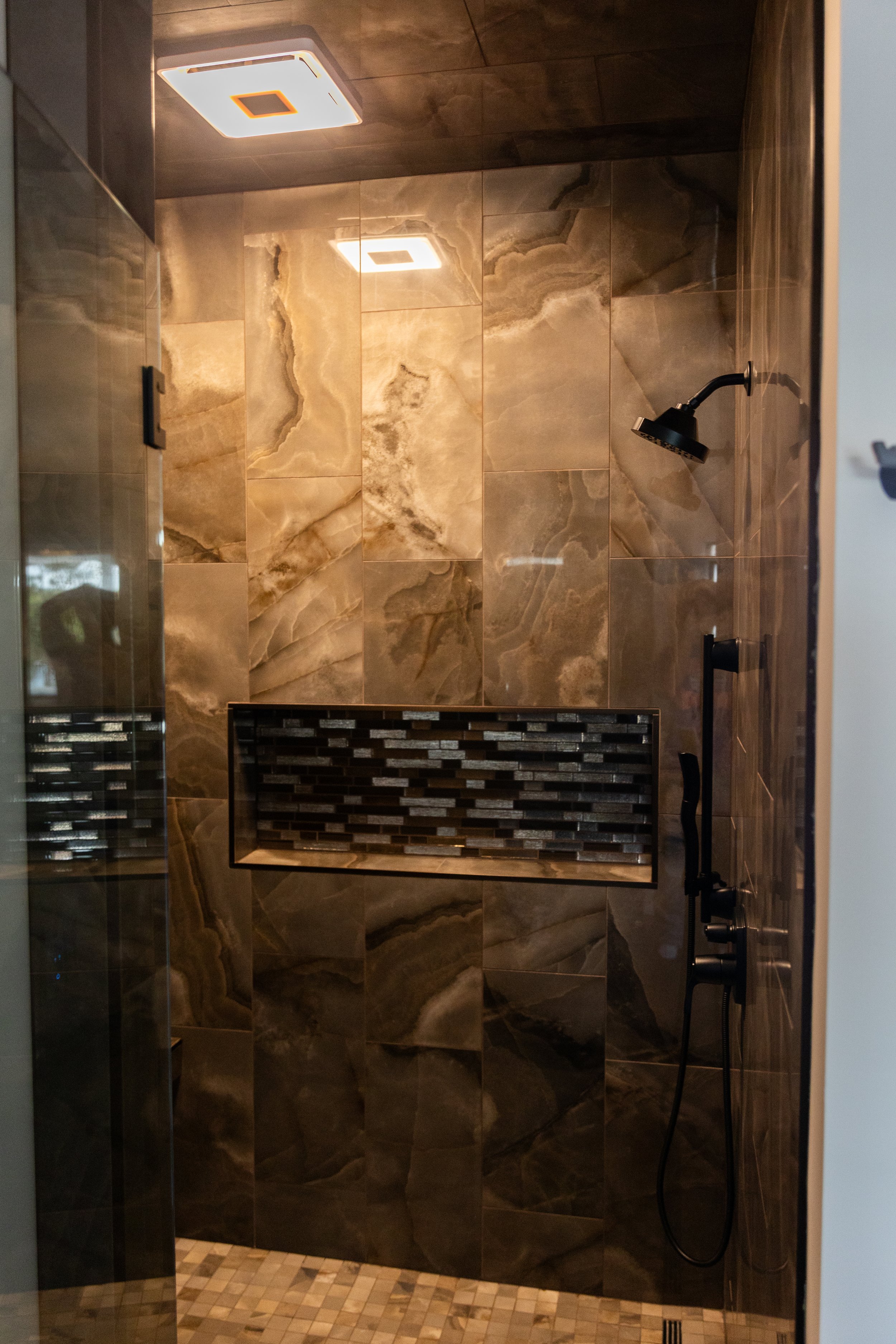 Shower area with dark marble tiles on walls, a black rainfall showerhead, black controls, a small window in the top corner, and a tiled floor with beige and brown mosaic stones.