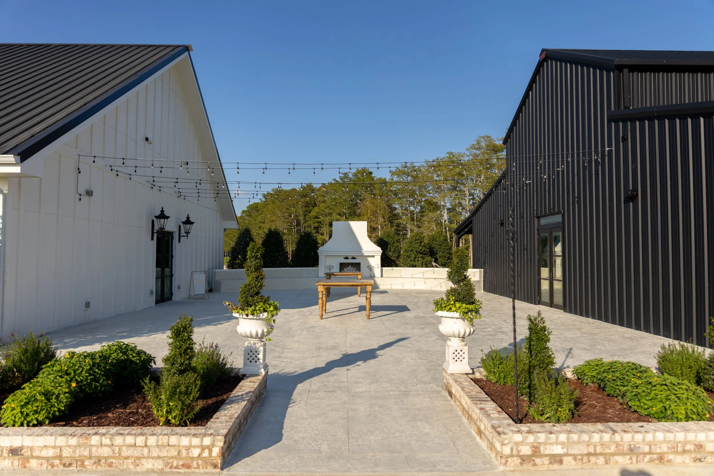 A landscaped outdoor patio area between two buildings, one painted white and the other black. The patio features potted plants, a table, string lights overhead, and a fireplace in the background.