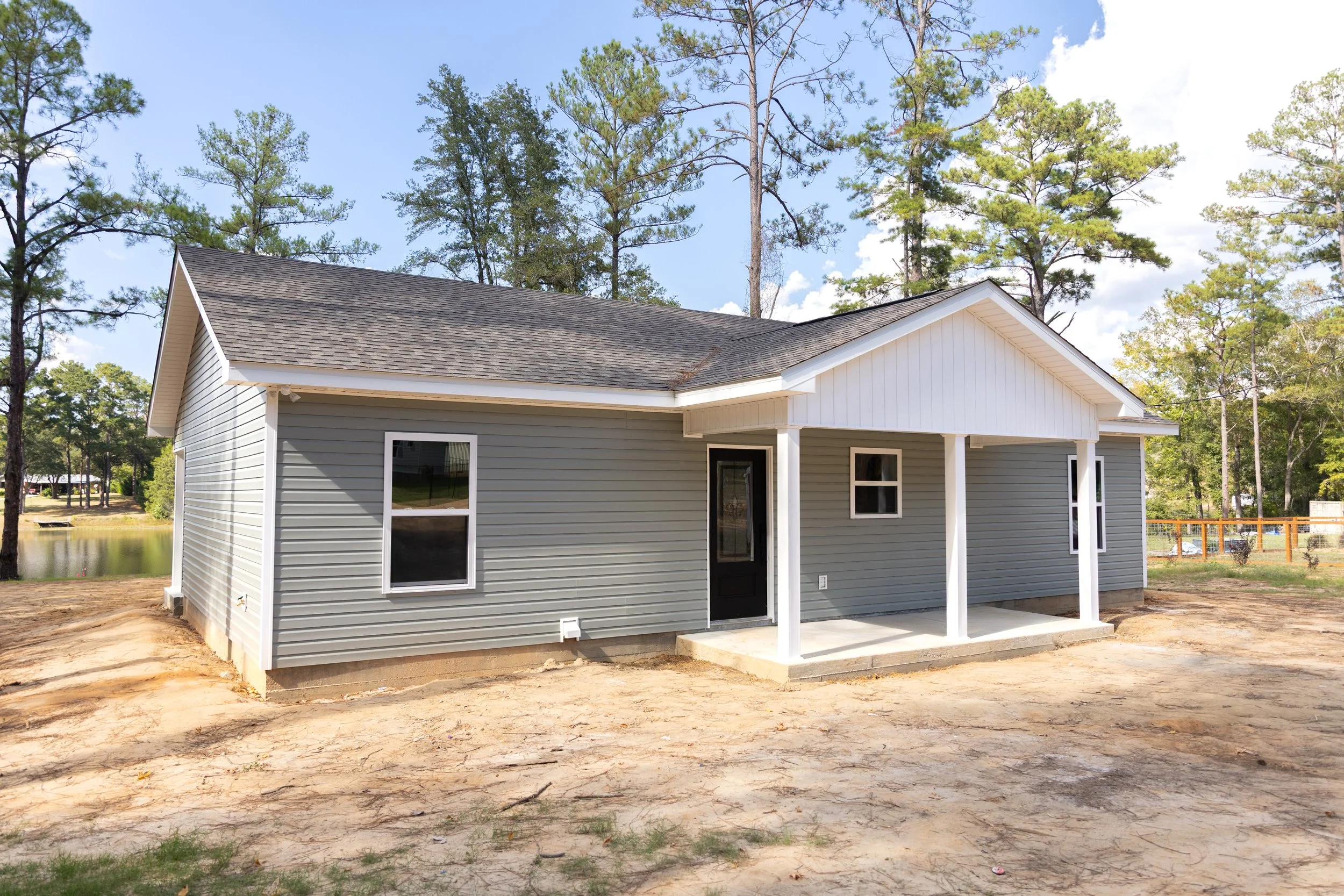 New gray house with white trim and a black front door, surrounded by trees, on a sunny day with a partly cloudy sky.