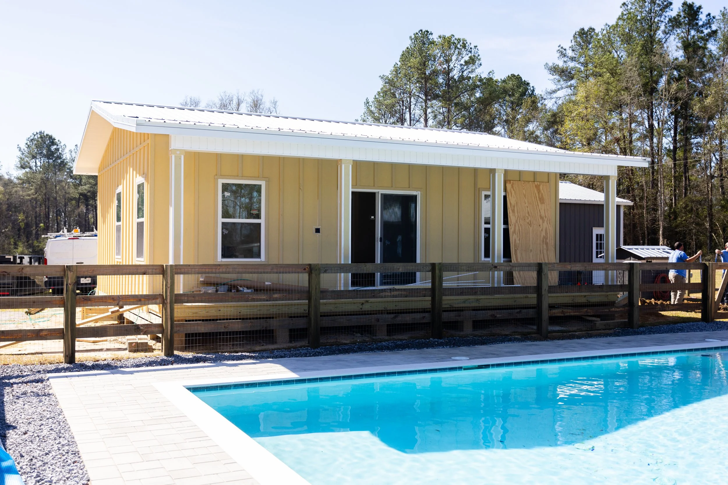 A residential house under construction with a swimming pool in the foreground, surrounded by a wooden fence, trees in the background, and construction workers visible.