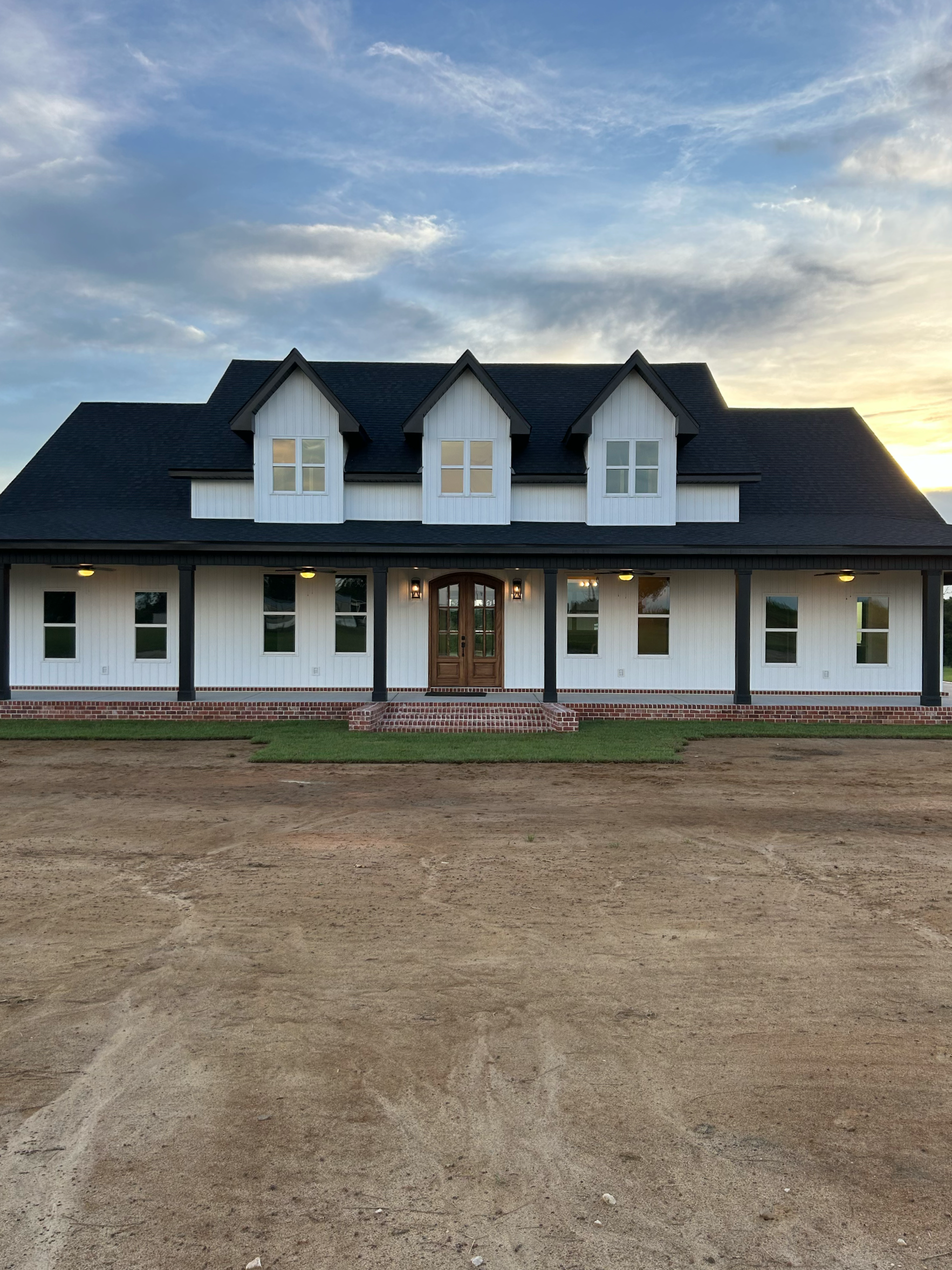 A two-story house with a dark roof, white exterior walls, multiple windows, and a brick porch with steps. The house is under a sky with scattered clouds at sunset or sunrise, and the foreground is an unpaved dirt area.