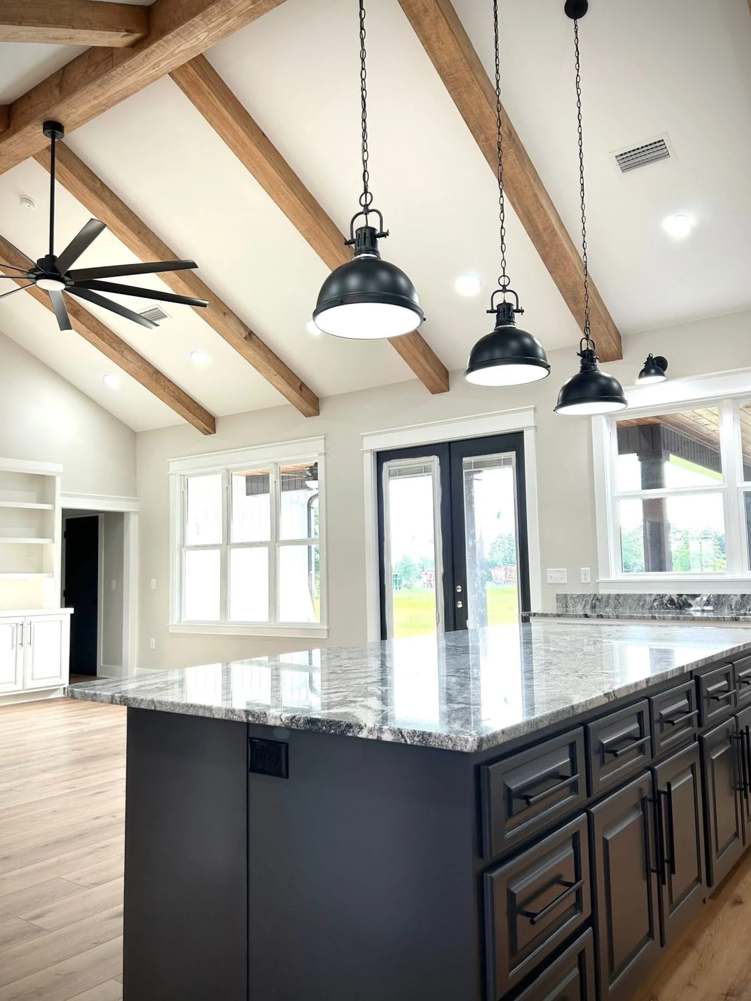 Modern kitchen with black cabinets, granite countertop island, wooden beamed ceiling, pendant and ceiling lights, and large windows.