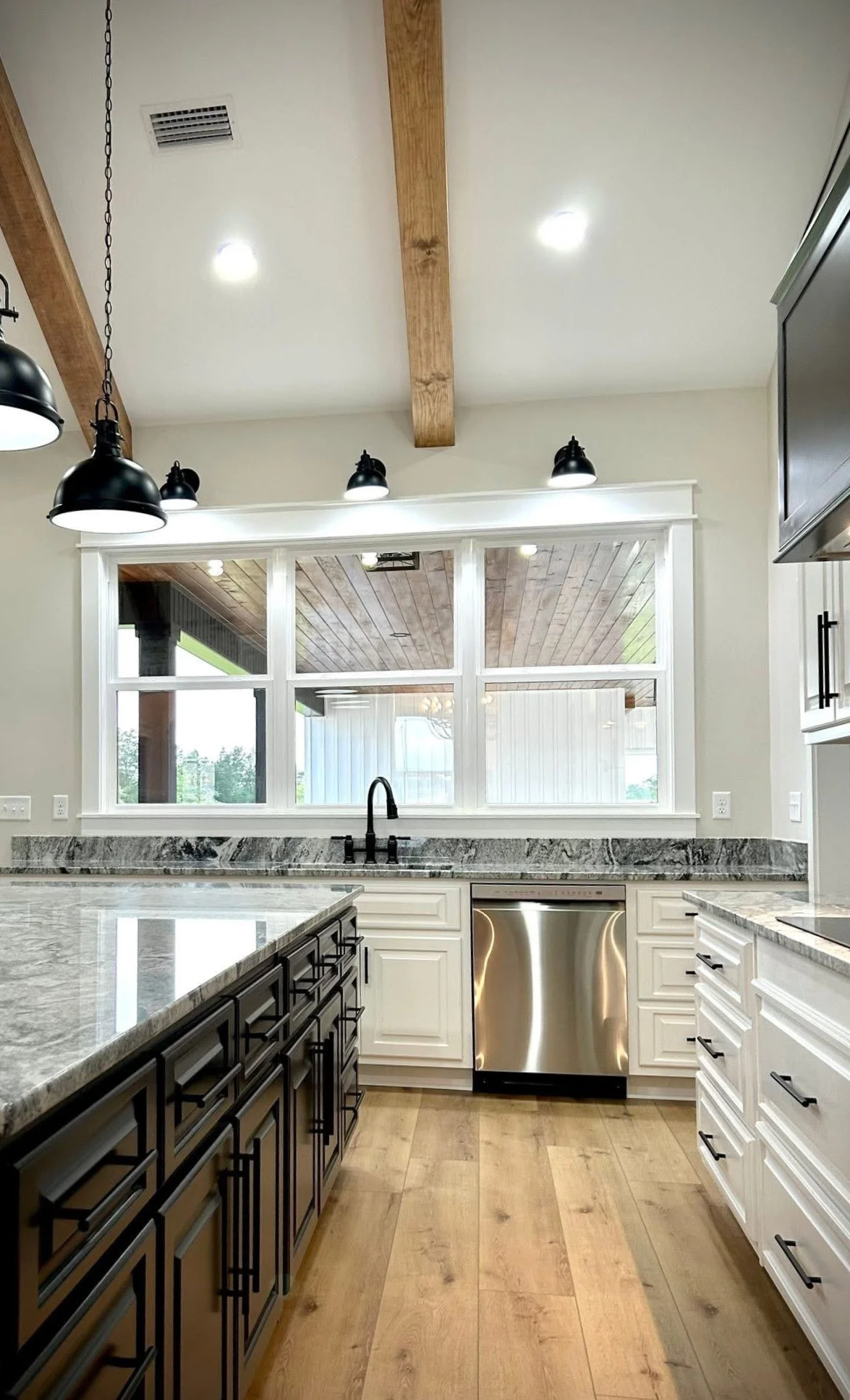 Modern kitchen with white cabinetry, black hardware, granite countertops, stainless steel dishwasher, black pendant and track lights, large window, and wooden ceiling beams.