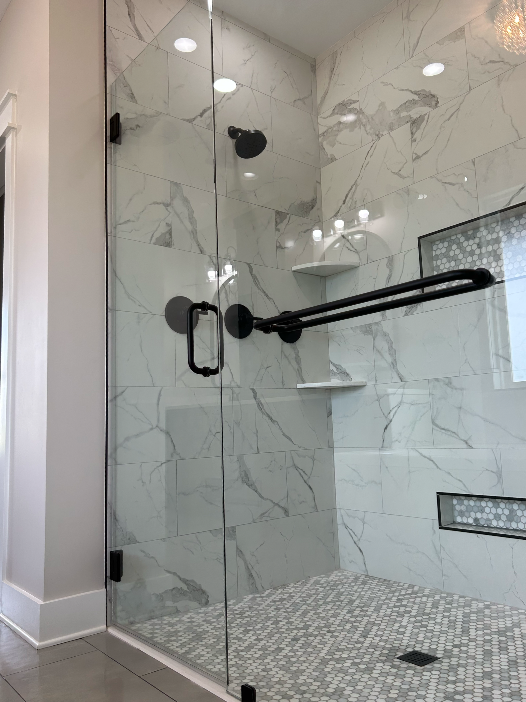 A modern glass-enclosed shower with marble tiles on the walls, black fixtures, and a white hexagon tile floor in a bathroom.