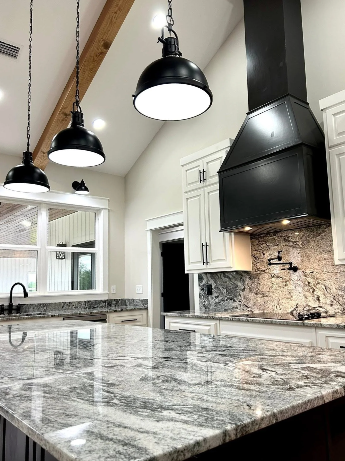 A modern kitchen with black pendant lights, white cabinets, and a granite countertop with a gray and white pattern. There's a black range hood above the stove, and a large window with a view outside.