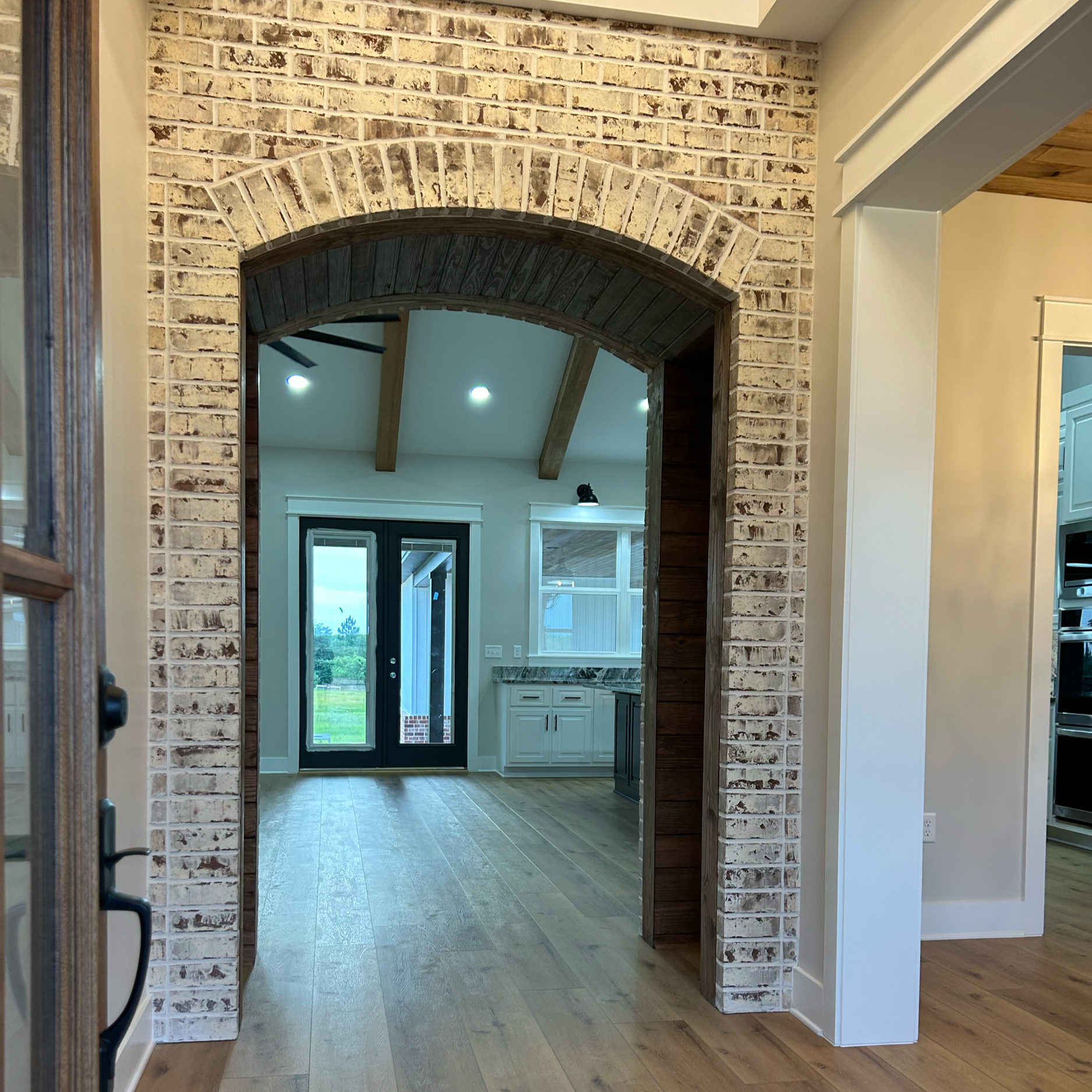 View of a kitchen entryway with brick arch framing a doorway leading to a room with large windows, wooden floors, and a ceiling with exposed beams, seen through a wooden frame.