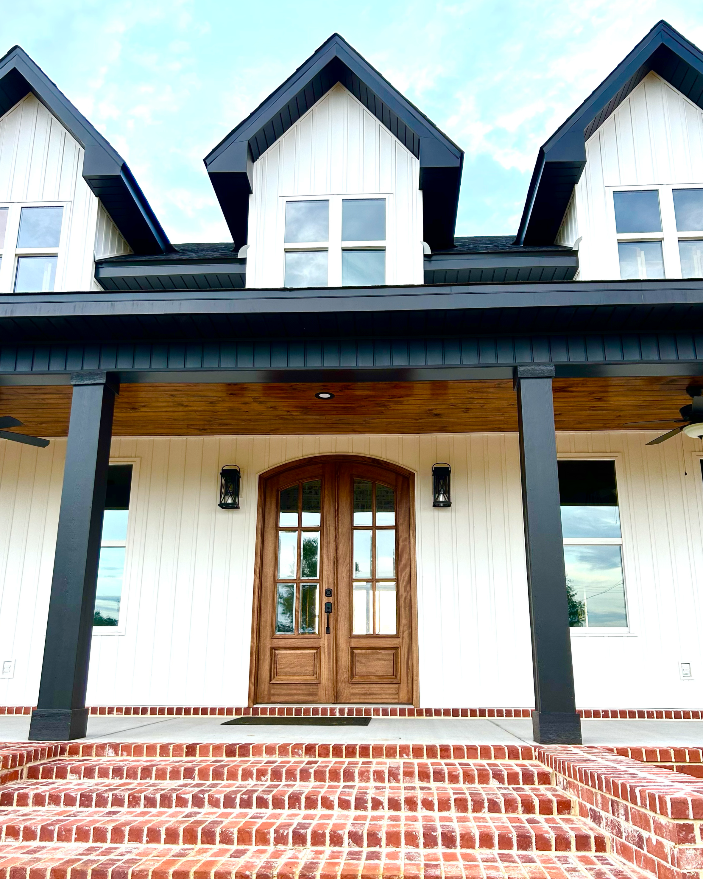 Front entrance of a house with a wooden double door, brick steps, black columns, and a porch ceiling, with a modern gabled roof with dormer windows and large windows on the facade.