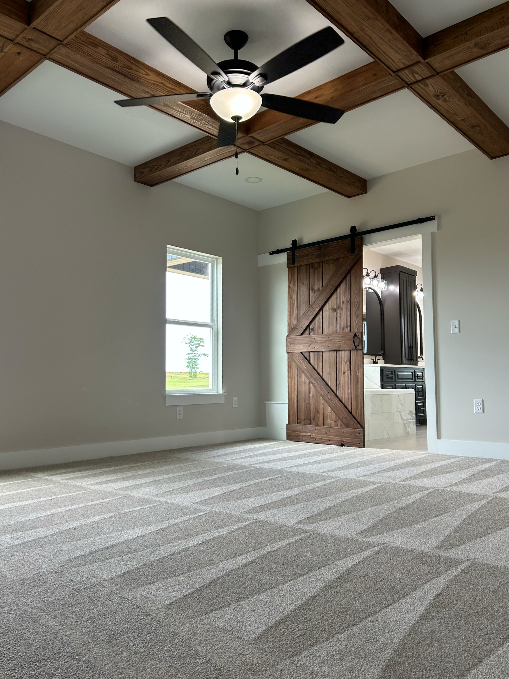 Living room with beige carpet, white walls, wooden ceiling beams, a ceiling fan with a light, a window letting in natural light, and a sliding barn door leading to a kitchen area.