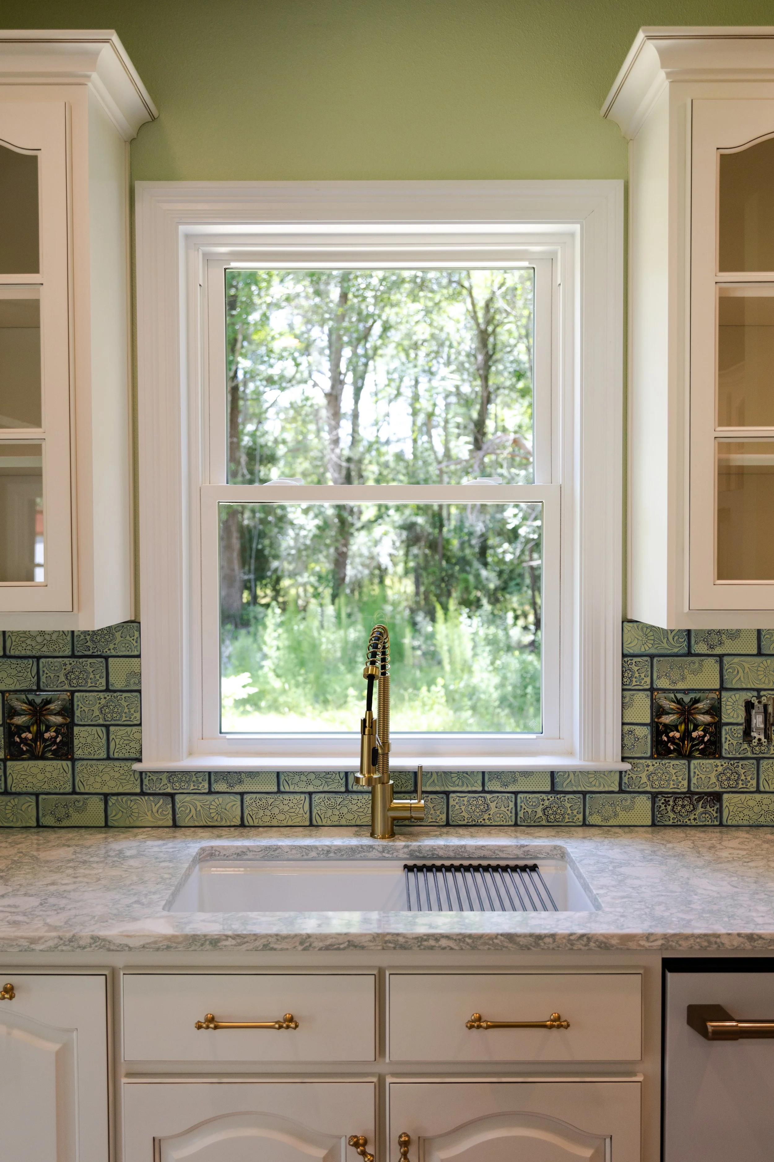 Kitchen sink with gold fixtures under a window with a view of greenery outside, tiled backsplash, and white cabinetry.