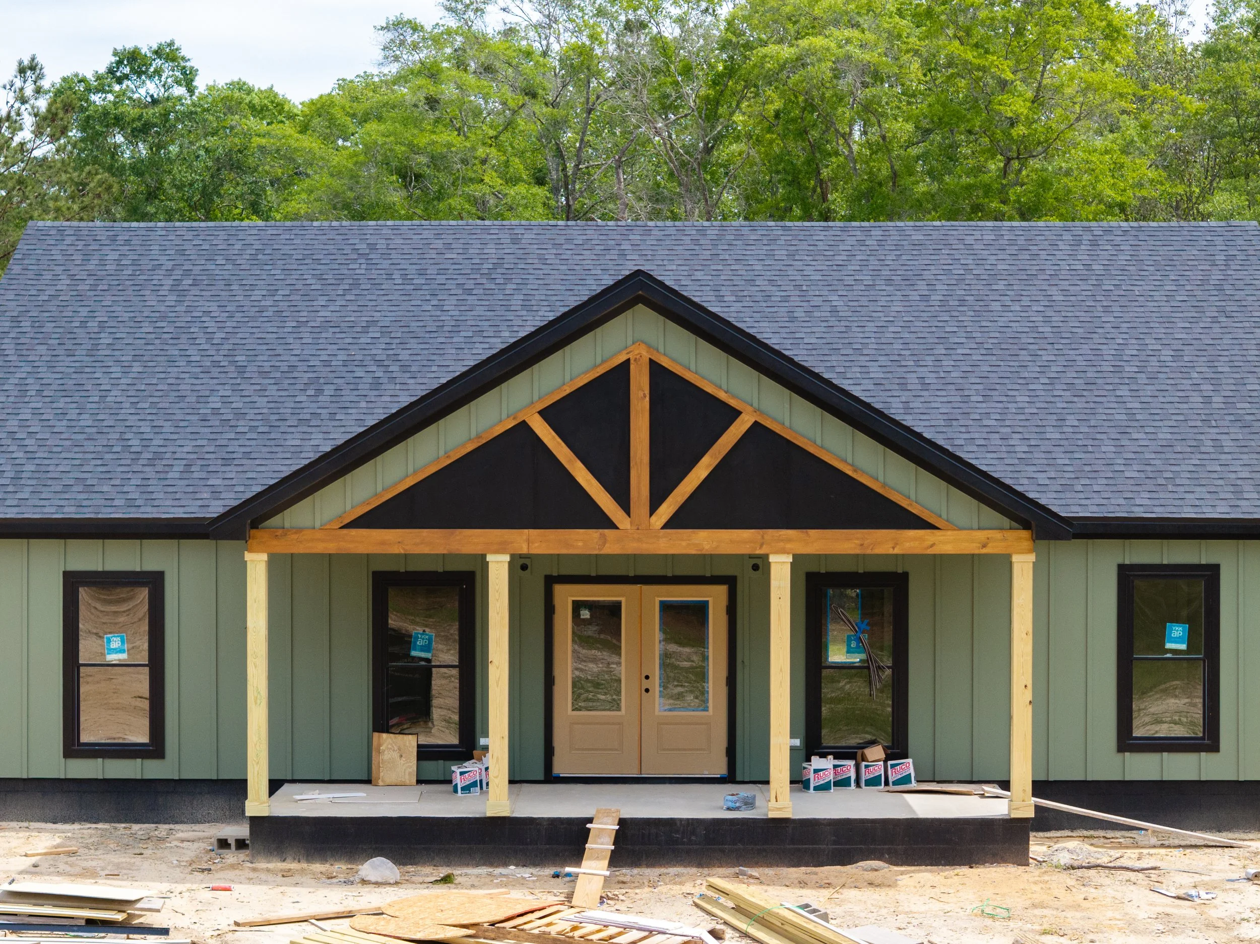 Front of a house under construction, with green siding, a gabled roof with gray shingles, and exposed wooden framing at the entrance, surrounded by construction materials and tools.