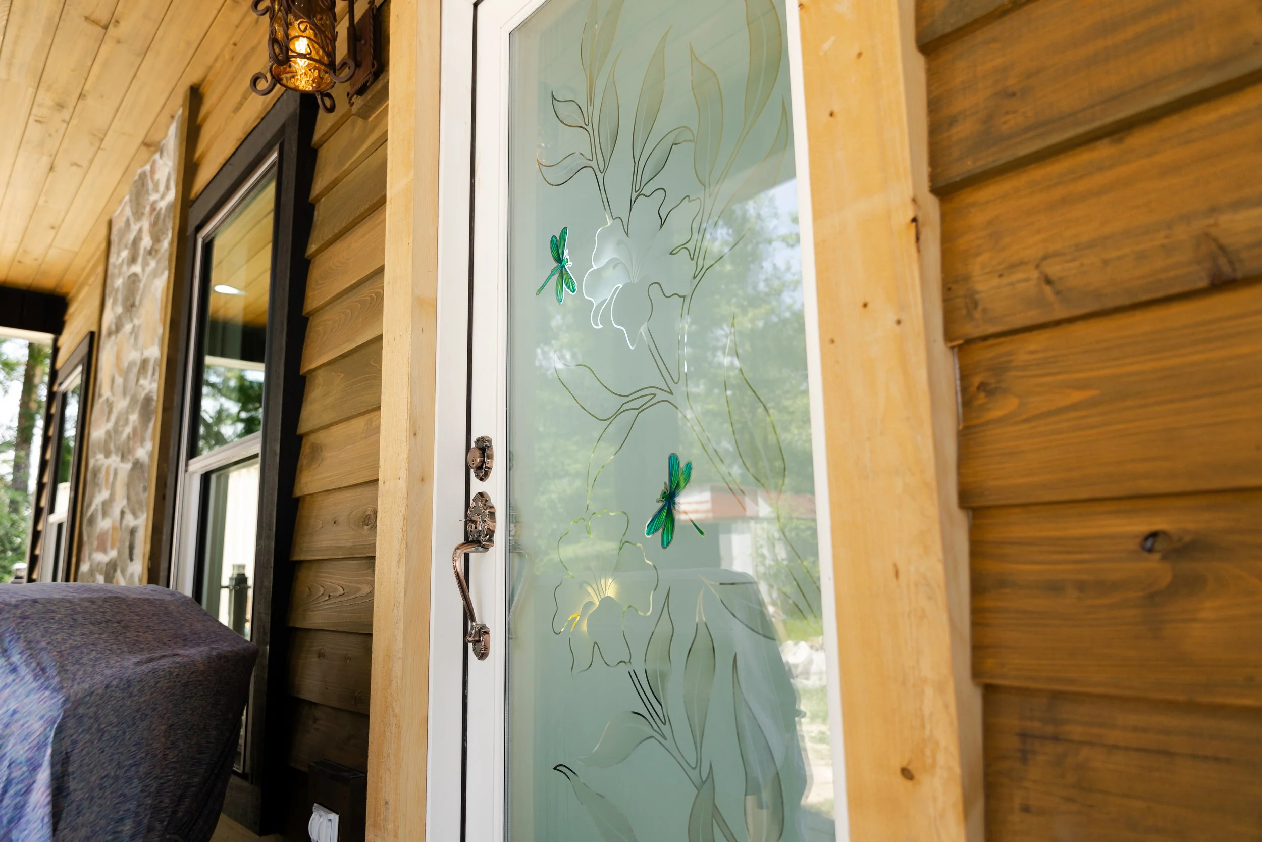 Close-up of a wooden porch wall with a large window decorated with dragonfly and floral stickers, a wrought iron outdoor sconce light, and a brass faucet below the window.